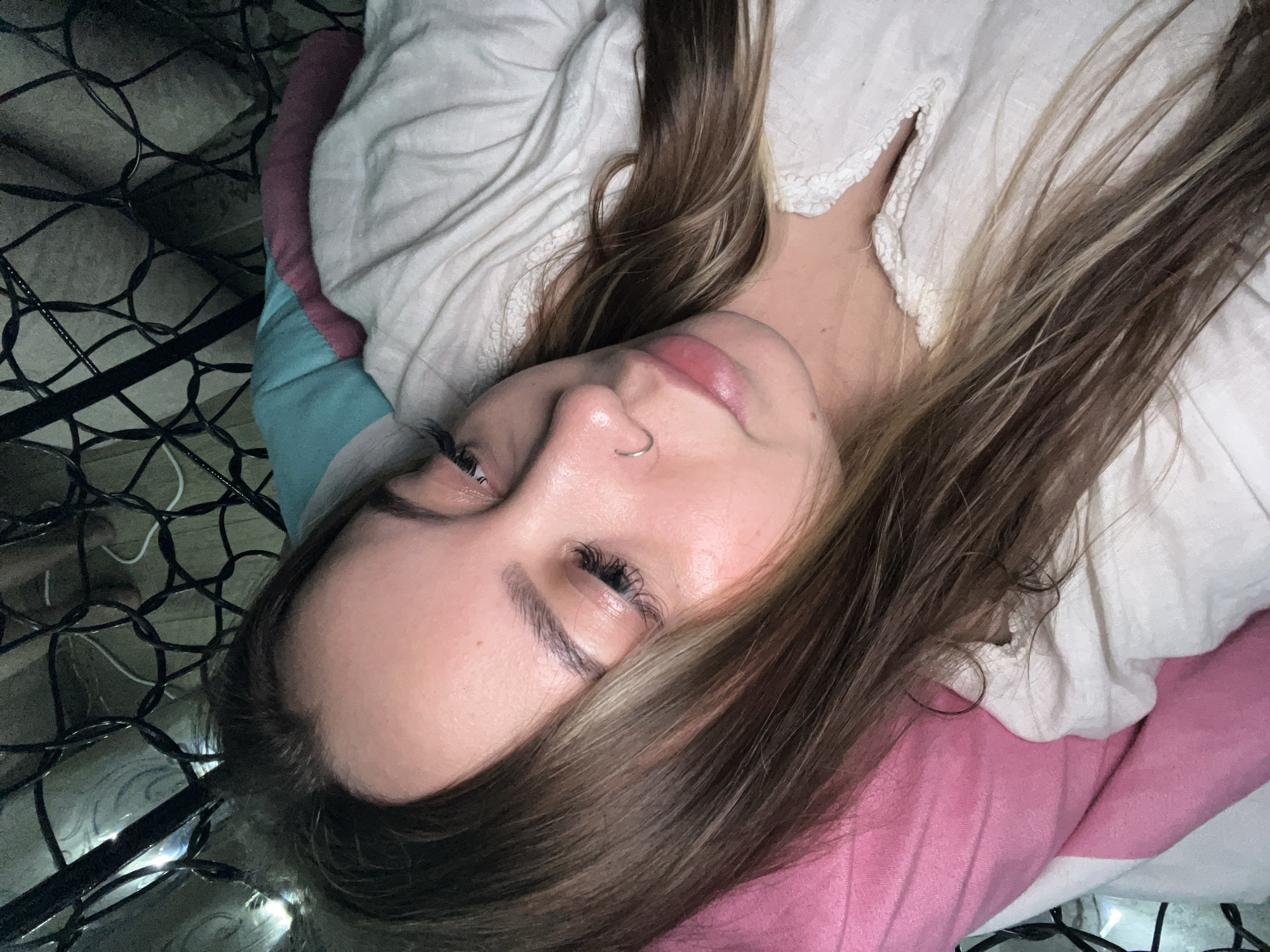 A person is lying down, viewed from above. The person has brown hair and fair skin, and is wearing a light-colored top. A nose ring is visible. The person is lying on a pink, white and blue pillow or cushion. To the left, there are black wires or string lights, possibly on a bed frame, and a glimpse of the floor. The location is unknown.