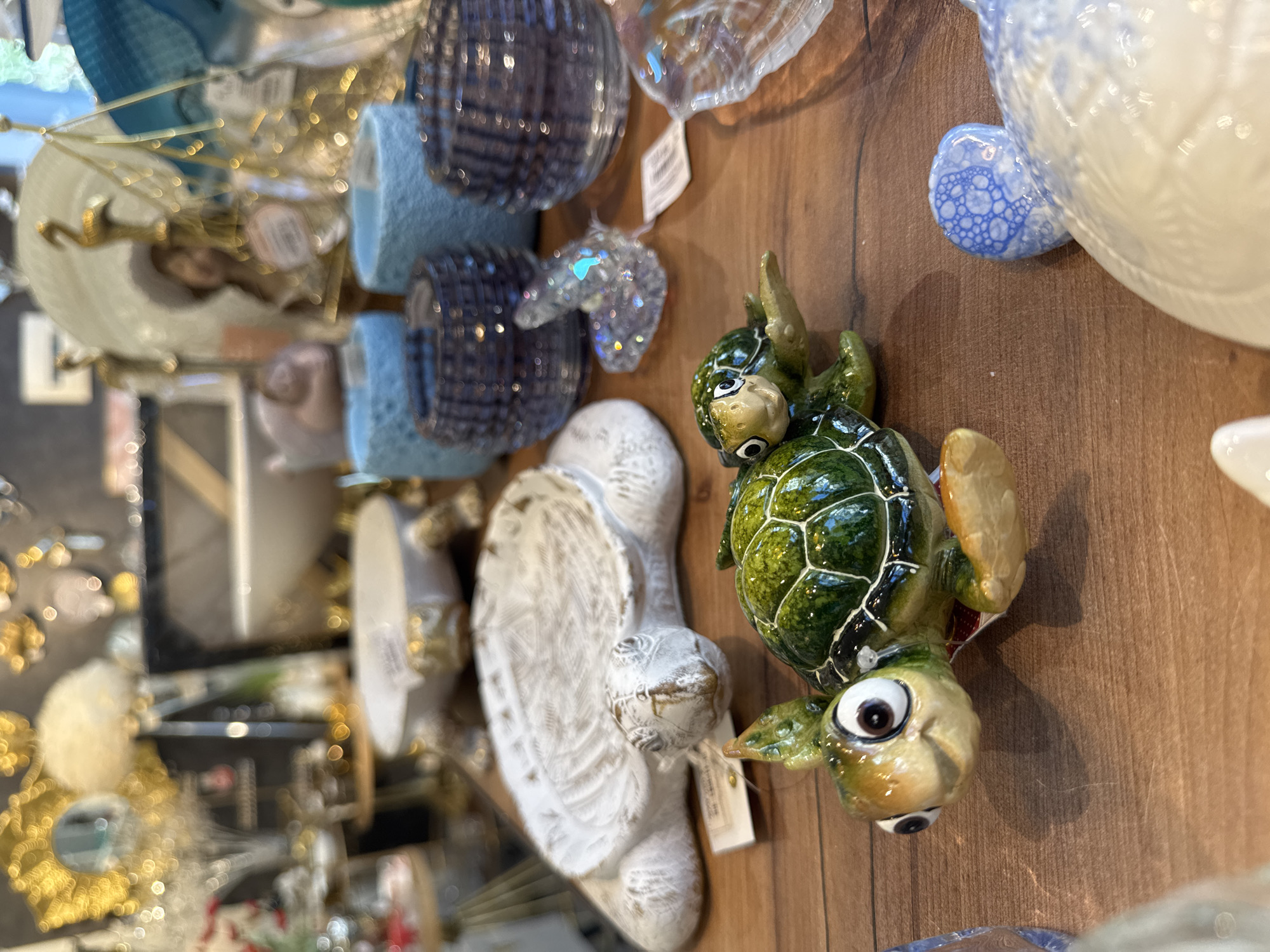 A close-up, eye-level shot captures decorative turtle figurines and other home decor items arranged on a wooden surface. In the foreground, a pair of glossy, green sea turtle figurines are prominently displayed. The larger turtle, positioned on the left, has large, expressive eyes and a slight smile, with a smaller turtle perched on its back. Behind them, a white ceramic turtle shell-shaped dish with gold accents rests on the wooden surface. To the right of the turtle figurines, there are several blue glass candle holders and a clear glass decorative item resembling a shell. In the background, out of focus, are various other decorative items, including gold-colored abstract sculptures and what appear to be decorative balls or spheres. The lighting is bright and slightly diffused, highlighting the textures and colors of the objects. There are no people visible in the image. The scene appears to be an indoor display in a retail setting, possibly a gift shop or home decor store, located in Sochi, Russia.