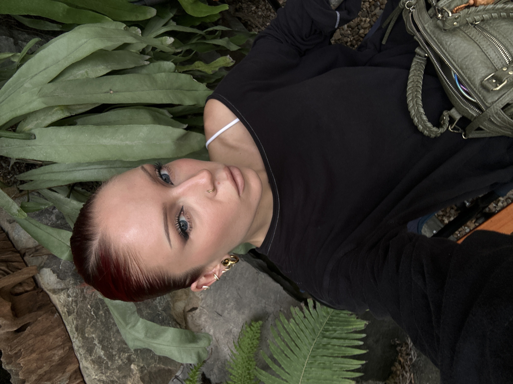 The image shows a close-up, over-the-shoulder selfie of a young adult with dark red hair pulled back, wearing a black long-sleeved shirt and a white bra strap visible. They have noticeable makeup, including winged eyeliner and lip gloss, and jewelry consisting of multiple ear piercings and a nose ring. They are holding a green, textured handbag. The background features large rocks and lush green foliage, possibly ferns, and appears to be an outdoor, natural setting. The lighting is soft and natural. There is no indication of the image being taken in Da Nang, Vietnam.