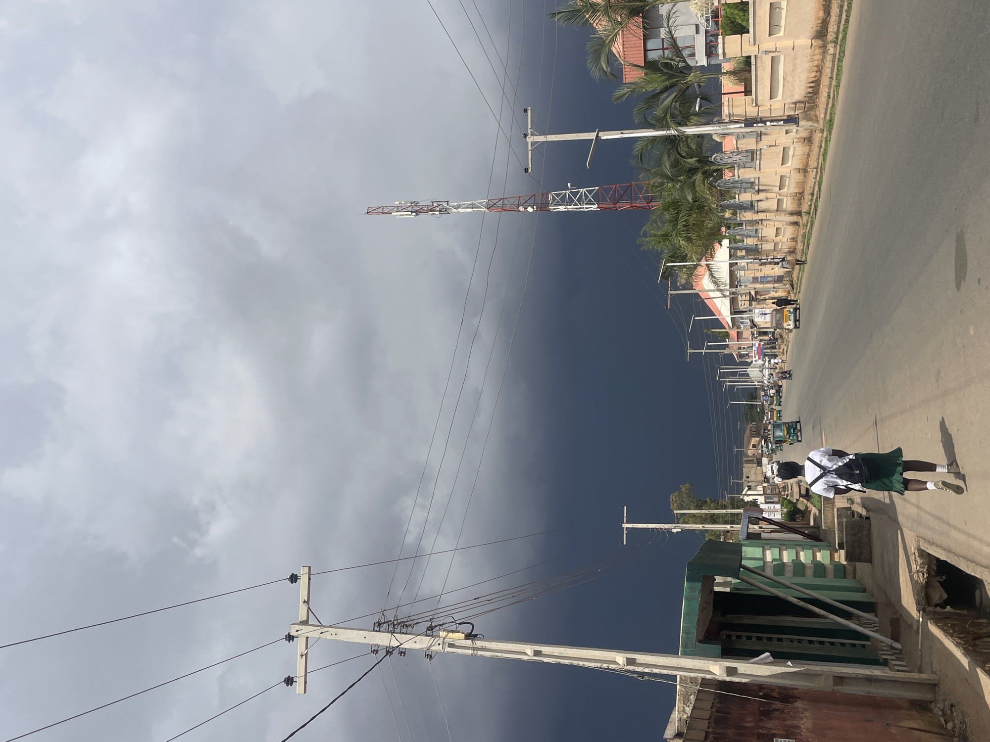 The image shows a street scene in Jos, Nigeria, under a dramatic sky split between light gray clouds and a dark blue mass. A tall red and white communication tower is visible in the distance. Along the street, there are buildings, utility poles with multiple wires, and vehicles. A person wearing a white shirt, dark skirt, and carrying a backpack walks along the right side of the street. A low building with green accents is in the foreground on the right.