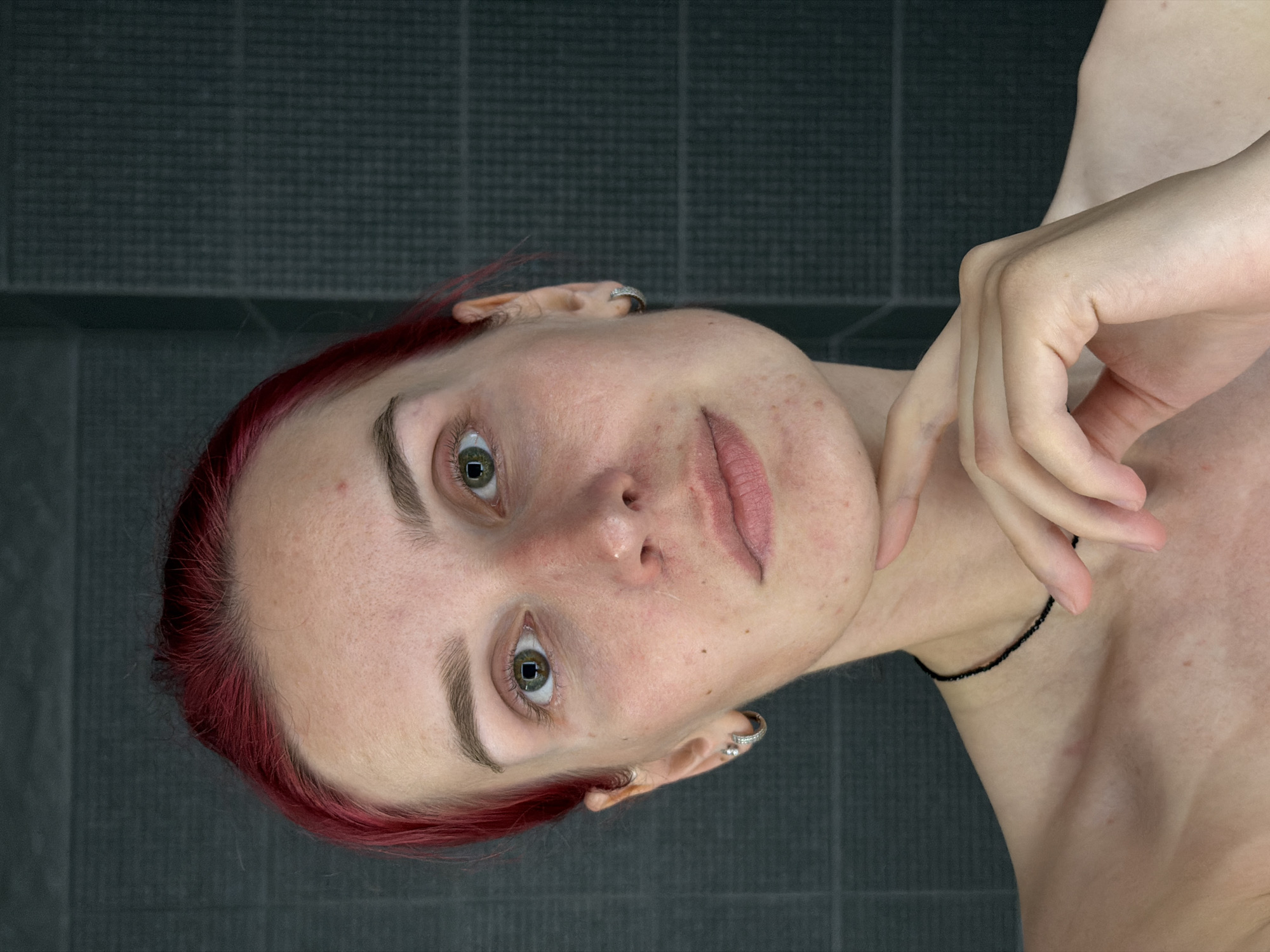 The image shows a close-up, front-facing portrait of a person with bright red hair, green eyes, and fair skin. The individual has a few blemishes and a small nose stud. They are wearing small hoop earrings and a thin black choker necklace. Their right hand is resting on their chin. The background appears to be a dark, tiled wall. There are no specific landmarks or elements in the image that would allow for the identification of a location within the city of Chaikovsky, Russia.