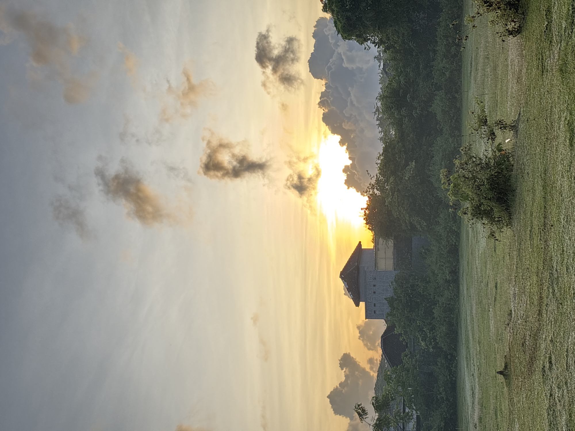 The image shows a landscape in Kuta Selatan, Indonesia, possibly near sunset or sunrise. A low-rise building or structure with a tiled roof is visible through trees. To the right of the structure is a green field. The sky is filled with clouds, illuminated by the sun. The sun itself is largely obscured by clouds, but the light is cast across the sky, creating a warm, golden glow.
