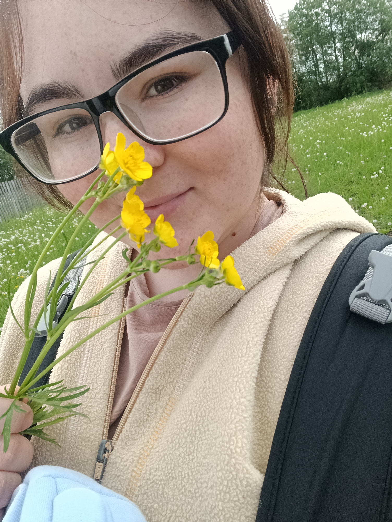 A woman with brown hair and glasses holds a bouquet of yellow wildflowers in front of her face. She wears a light-colored fleece jacket and a backpack with a black strap is visible over her shoulder. The background shows a grassy field dotted with white flowers, with trees visible in the distance. The location is outdoors, likely in a rural or natural setting. The city of Jelpachiha, Russia is not discernible from the information given within the scene.