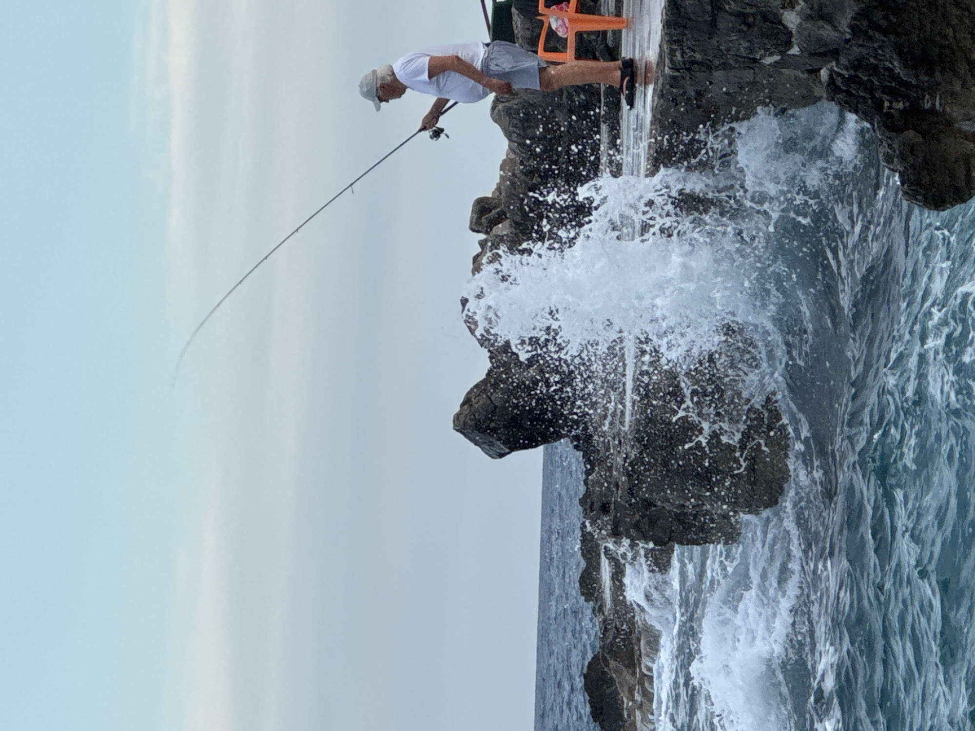A man is fishing on a rocky shore in Prijevor, Montenegro. He is standing on a concrete walkway, holding a fishing rod with his right hand. He is wearing a white t-shirt, grey shorts, a white cap, and dark sandals. A wave is crashing against the rocks in front of him, sending spray into the air. The sea is dark blue and choppy. In the background, there is an orange plastic chair. The sky is overcast.