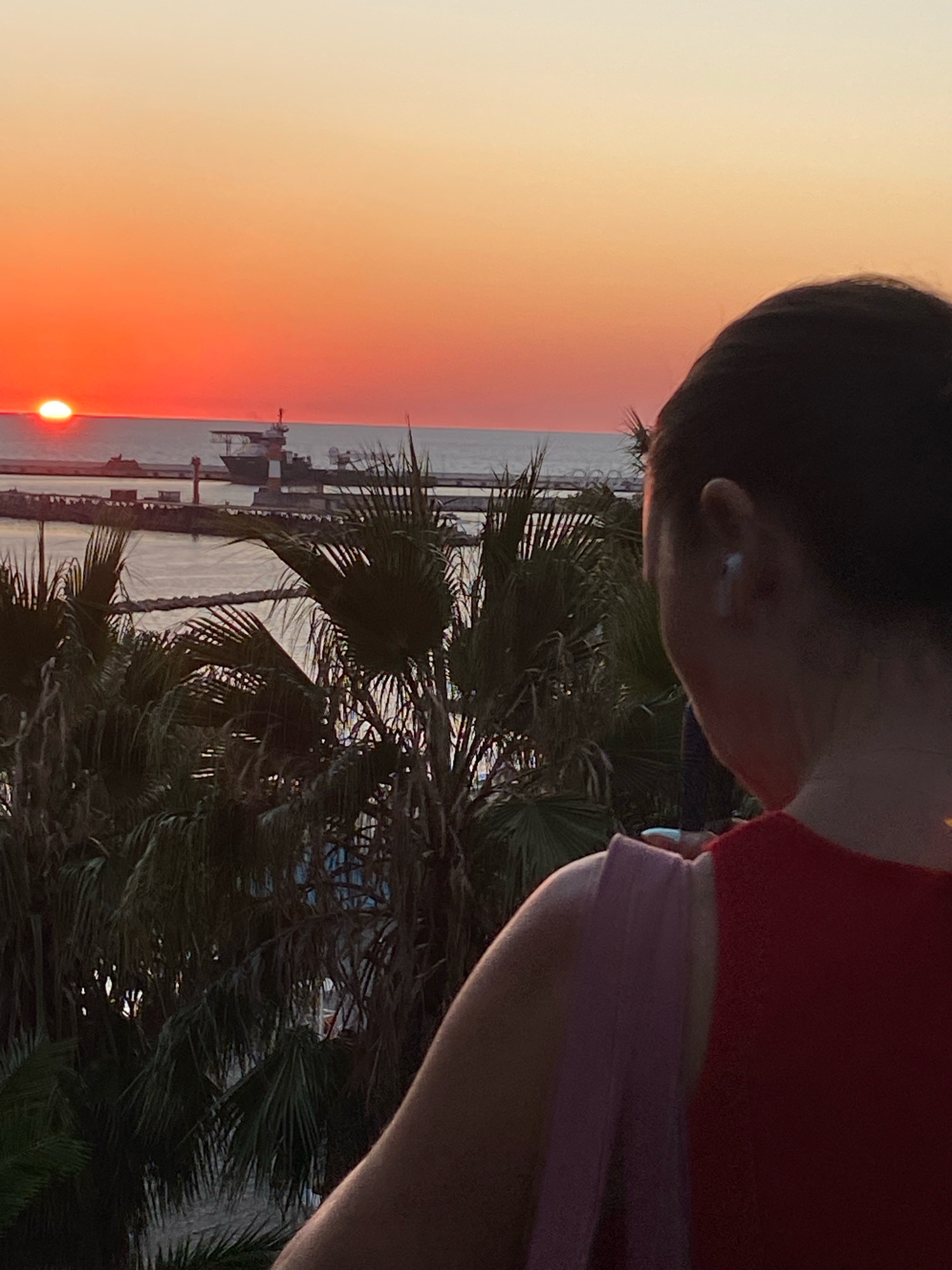 A woman is seen from behind, looking out at a sunset over the sea. She has dark hair pulled back and is wearing a red top with a pink strap over her shoulder. She has a white earbud in her right ear. The sun, a bright orange orb, is low on the horizon, casting a warm glow across the sky and sea. In the distance, a cargo ship is visible docked at a port with breakwaters. Palm trees frame the foreground, their fronds dark against the vibrant sunset. The scene appears to be from a balcony or elevated viewpoint, likely in a coastal city such as Sochi, Russia, known for its Black Sea coastline and palm trees.