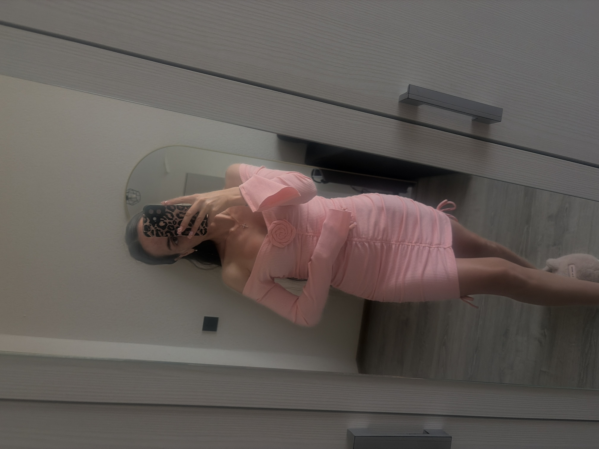 A young woman, whose face is obscured by a phone, stands in front of a full-length mirror, taking a selfie. She is wearing a light pink, off-the-shoulder, long-sleeved mini dress with ruching down the sides and a rose embellishment on the bust. The dress has tie details on the sides. She is also wearing fuzzy slippers. The background appears to be a bedroom or dressing room, with a closet door to her right and a full-length mirror to her left. The flooring is a wood-grain laminate. The lighting is soft and diffused.