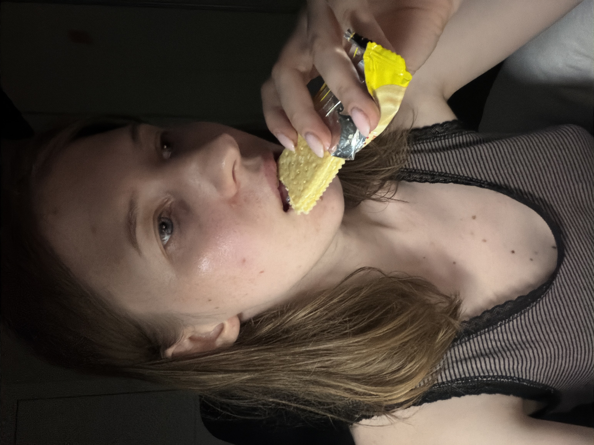A young Caucasian woman with light brown hair, wearing a black and gray striped tank top, is captured in a close-up shot as she eats a cracker from a yellow wrapper. Her right hand holds the cracker near her mouth, and her left hand is visible near the wrapper. The background is dark, suggesting an indoor setting, possibly at night. The lighting highlights her face, revealing a fair complexion with some minor skin imperfections and a sheen of sweat. The image appears to be a candid, personal photograph. No specific location in Dongducheon-si, South Korea can be determined from the visual information provided.