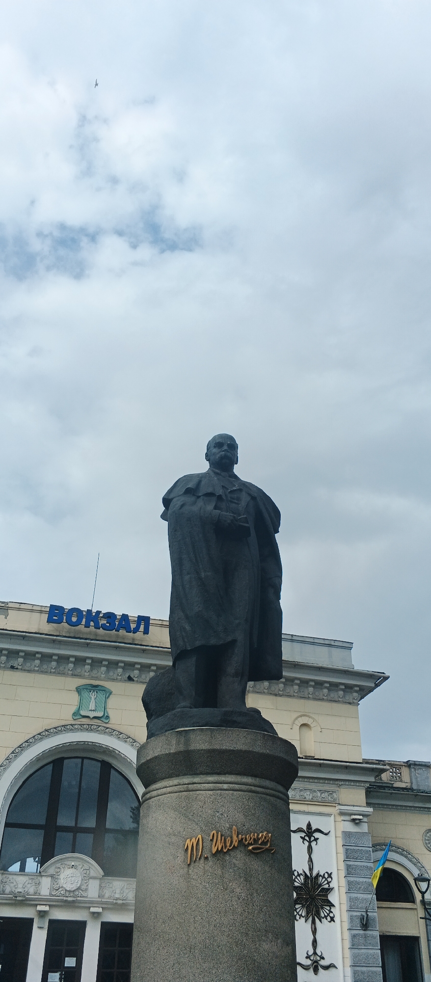 In Smila, Ukraine, a dark bronze statue of a man stands prominently in front of a cream-colored building with blue lettering that spells "ВОКЗАЛ" (Vokzal, meaning "station"). The statue depicts a figure in a long coat, holding something in his hand, and is mounted on a stone pedestal. The pedestal has a golden inscription that appears to read "М. Шевченко" (M. Shevchenko). The building features arched windows and decorative architectural elements. A Ukrainian flag is visible on the right side of the building. The sky above is overcast with clouds, and a small airplane is visible in the upper left quadrant of the image.