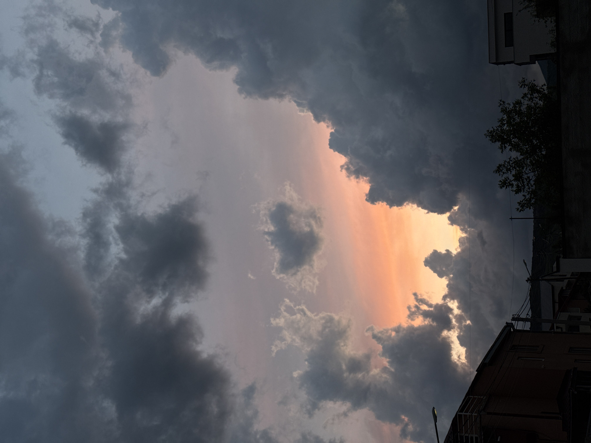 The image depicts a dramatic sunset sky over a residential area of Tbilisi, Georgia, characterized by heavy cloud cover and distinct architectural elements. The majority of the sky is filled with dense, dark grey storm clouds, some showing texture and volume. A prominent opening in the cloud mass, towards the bottom-center and right, reveals a vibrant sunset glow, transitioning from intense orange at the horizon to softer pinks and purples higher up, against a backdrop of lighter grey and pale blue sky in the upper left quadrant.
In the lower portion of the frame, several buildings and urban infrastructure elements are visible in silhouette or low light. On the left, a two-story building with a reddish-brown facade features a white metal railing on an upper balcony or terrace. A bright streetlamp is illuminated and visible above this building. Towards the center and right, multiple utility poles support a network of overhead power lines that traverse horizontally across the midground. A second, less prominent streetlamp is visible to the right. On the far right, a segment of a modern, light-colored (possibly white) building with a dark, flat roofline is partially visible, partially obscured by a silhouetted deciduous tree with dense foliage. Distant, dark hills or mountains form an indistinct horizon line in the far background. No people, animals, or vehicles are discernible in the scene. The overall lighting suggests dusk or early evening, intensified by the dramatic interplay of storm clouds and sunset colors.