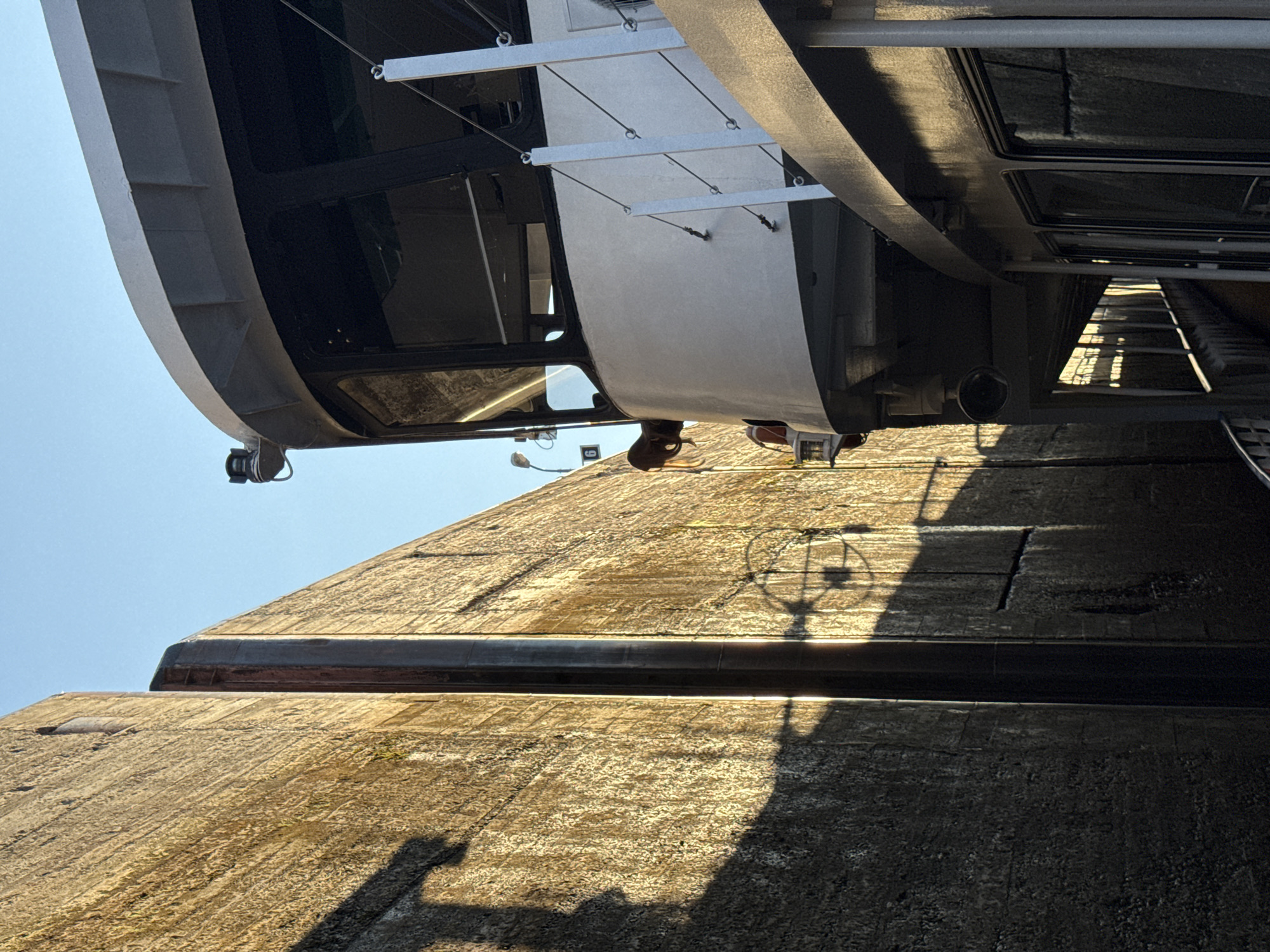 An extreme close-up shot captures a section of a canal lock wall and a portion of a white passenger boat. The canal wall, a rough concrete structure, is illuminated by warm, angled sunlight, casting long shadows. A dark, vertical metal beam runs along the seam where two sections of the wall meet. The boat, positioned to the right, has a white superstructure with dark-tinted windows. A white railing with wire supports is visible along its deck. The sky above is a clear, bright blue. The scene depicts the boat navigating through the narrow confines of the lock.