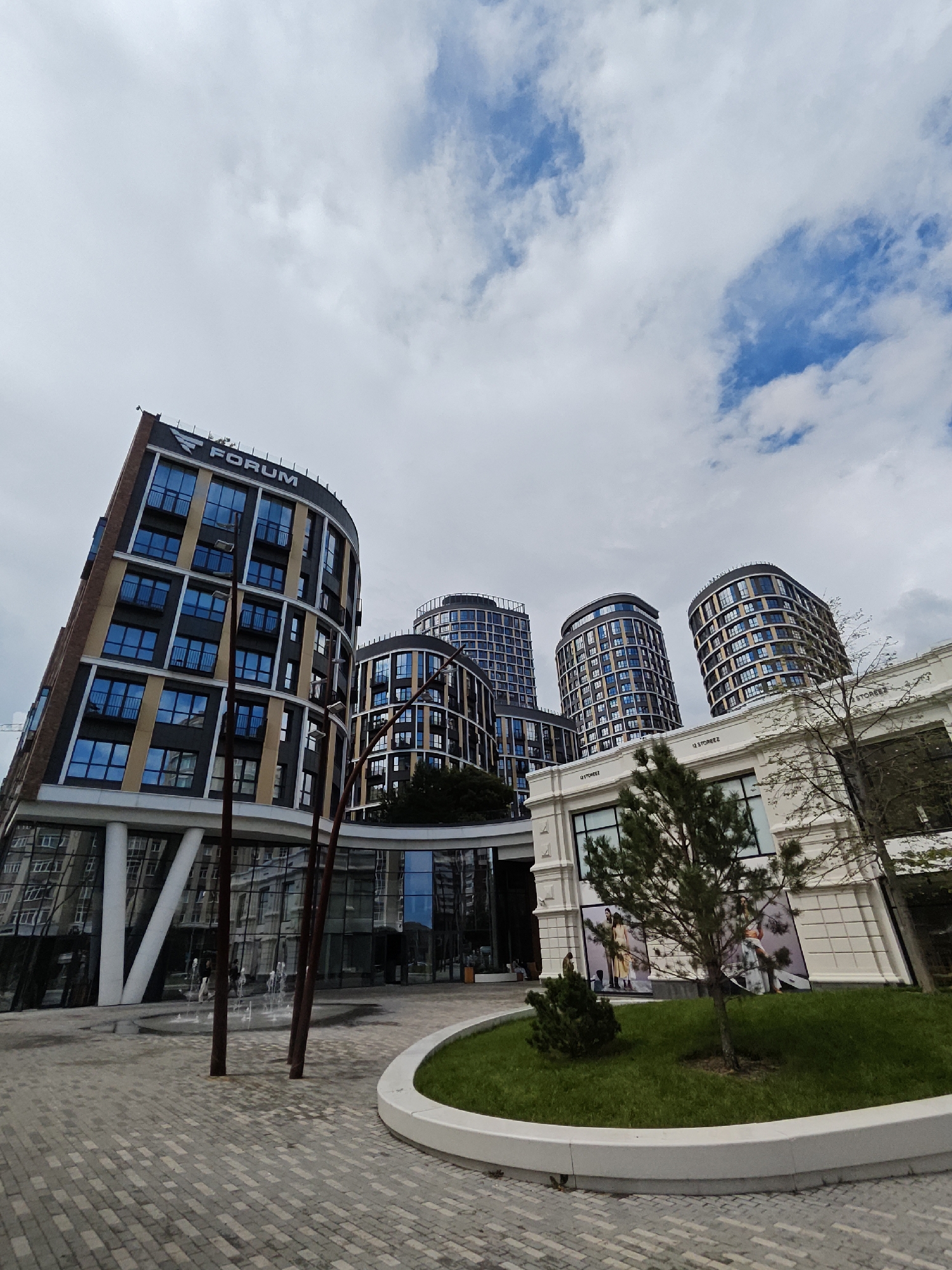The image depicts a modern urban scene featuring contemporary architecture. In the foreground, a paved plaza with a curved white border leads to a circular patch of green grass with a young tree. Several dark, vertical posts stand in the plaza, near a small fountain with water jets. Behind the fountain, a building with a glass facade reflects the surrounding structures.
Dominating the midground and background are several tall, curved apartment buildings. The building on the left is a prominent dark structure with horizontal bands of brown and light yellow, and numerous blue-tinted windows. It features the word "FORUM" in large white letters at the top. To its right, a cluster of similarly curved, multi-story buildings with dark facades and grid-like patterns of windows are visible.
In the right side of the midground, a white, modern building with large windows and a tiled facade is present. Large advertisements featuring people in clothing are displayed on the facade of this white building. The sky above is partially cloudy, with patches of blue visible. The image captures a well-maintained, contemporary urban development, likely a residential or mixed-use complex. This is the "Forum" residential complex in Yekaterinburg, Russia.