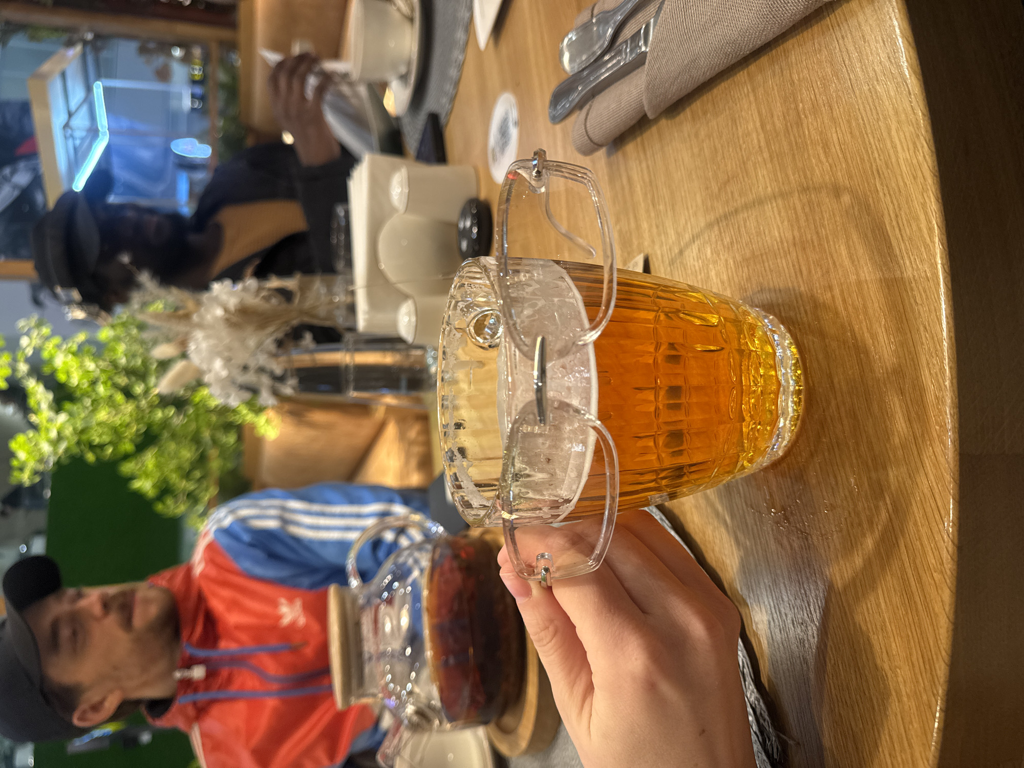 An indoor scene, likely a cafe or restaurant in Ul'yanovka, Russia, shows people gathered around a wooden table. The image is taken from a first-person perspective, focusing on a person's hand holding a unique glass mug filled with a clear, amber-colored beverage, possibly tea or juice. The mug features a distinctive lid with a hinged handle mechanism.
To the left, a man is visible wearing a red and blue Adidas track jacket with the brand's logo clearly displayed on his chest, and a black baseball cap. He appears to be looking towards the camera or another person at the table. Next to him, a clear glass teapot, also containing a dark liquid, rests on the table. In the background, further left, another person in dark clothing is partially visible, seemingly engaged with a phone or menu.
The table, made of light-colored wood with visible grain, is set with cutlery wrapped in a light napkin, white ceramic cups, and saucers. Decorative elements such as green foliage and dried ornamental plants are also present on the table. The lighting is warm and artificial, suggesting an evening or indoor setting. Reflections on the table surface indicate a glossy finish or slight dampness. The overall atmosphere appears casual and relaxed, with individuals enjoying drinks.