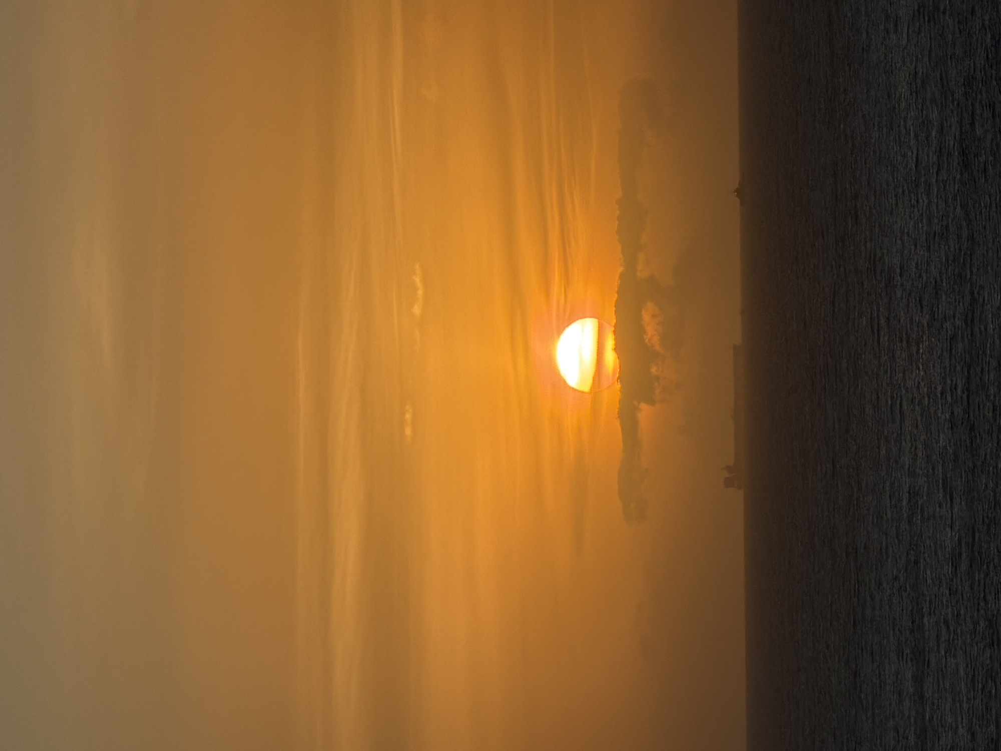 Captured from an elevated vantage point, this image showcases a partial sun obscured by clouds and atmospheric haze, with a body of water filling the scene to the right. The sun appears as a bright yellow orb, partially concealed by a dark, cloud-like formation. Streaks and patches of atmospheric haze diffuse the sunlight, creating an orange and yellow gradient. The body of water extends horizontally, contrasting with the light of the sunset. There are some buildings present on the edge of the horizon line. The image is oriented sideways, causing the horizon to appear vertical, but the primary focus remains the atmospheric conditions and light. The location within the city of Unknown, Unknown, is not clear and cannot be determined from this image alone.