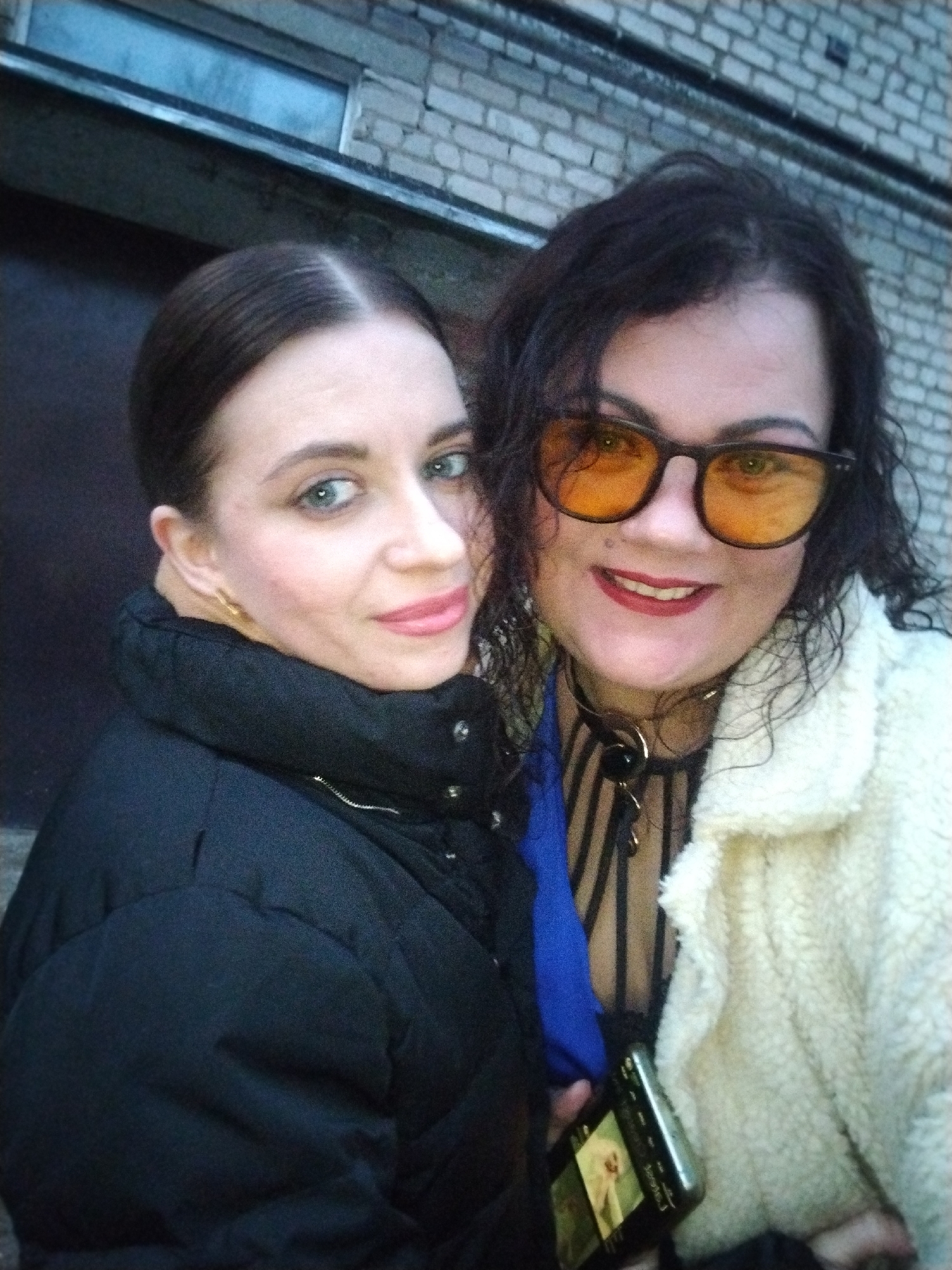 Two women are featured in a close-up, casual photograph, likely a selfie, taken outdoors against a brick wall backdrop, characteristic of an urban or suburban setting in Chaikovsky, Russia.
The woman on the left has dark hair styled neatly back, revealing light blue-green eyes and a fair complexion with a hint of pink blush. She wears pink lipstick and a gold earring. Her attire consists of a black puffer jacket with a high, protective collar, and she offers a subtle smile towards the camera.
The woman on the right has voluminous dark, curly hair and is wearing orange-tinted sunglasses with dark frames. Her lips are adorned with red lipstick, and she smiles broadly. She is dressed in a cream-colored, fluffy or sherpa-style jacket. Beneath it, glimpses of a blue garment and a black-and-white striped item are visible. A black choker-style necklace with a prominent gold-colored, spiral-like pendant is around her neck. She holds a smartphone, which displays an image on its screen, appearing to be a picture of a blonde woman.
The lighting suggests natural daylight, possibly overcast, as there are no harsh shadows. Both subjects appear happy and relaxed, exuding a friendly atmosphere. The brick wall behind them is a uniform pattern, providing a neutral background. Their jackets indicate cooler weather conditions. There is no visible text in the image itself, only the visual content on the smartphone screen.