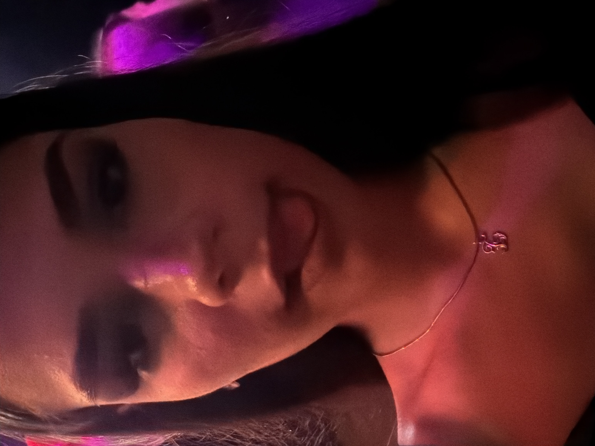 A close-up portrait photograph captures a person, likely female, from the chest up, oriented with her head towards the left side of the frame. Her face is turned partially towards the viewer, with her left eye, nose, and the side of her mouth visible. She has dark hair, which is illuminated by a strong purple light from above, creating vibrant streaks and reflections. Her skin, particularly on her face and neck, is bathed in a warm, reddish-orange light, creating contrasting shadows and highlights.
The subject appears to be mid-expression, with her tongue slightly extended in a playful manner. Her left eye shows visible dark makeup, possibly eyeliner or eyeshadow. Around her neck, she wears a delicate gold-toned chain necklace with a small, intricate pendant that resembles a stylized letter 'S' or a musical symbol, resting just above her collarbone.
The background is dark and out of focus, suggesting an indoor setting with artificial atmospheric lighting, such as a nightclub, bar, or a social event. The combination of intense purple and reddish-orange lighting indicates the photograph was taken at night. The overall mood is intimate and energetic. The location context provided is Varna, Bulgaria. No visible text is present in the image.