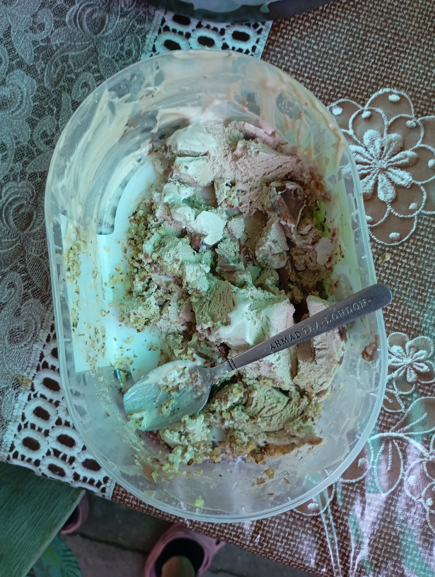An overhead, slightly angled close-up shot shows a partially eaten container of ice cream. The container appears to be clear plastic and is filled with a mixture of light brown and pale green ice cream, with visible chunks of nuts and possibly chocolate. A silver spoon rests on top of the ice cream, with the engraving "AHMAD TEA - LONDON" visible on its handle. The container sits on a patterned placemat. The surrounding environment suggests an indoor setting, likely a kitchen or dining area, with hints of furniture or other household items visible in the periphery, but no discernible cityscape or specific location within Horishni Plavni, Ukraine can be identified. There are no people clearly visible in the frame, only glimpses of feet in pink slippers at the bottom of the image. The lighting is somewhat dim and appears to be artificial.