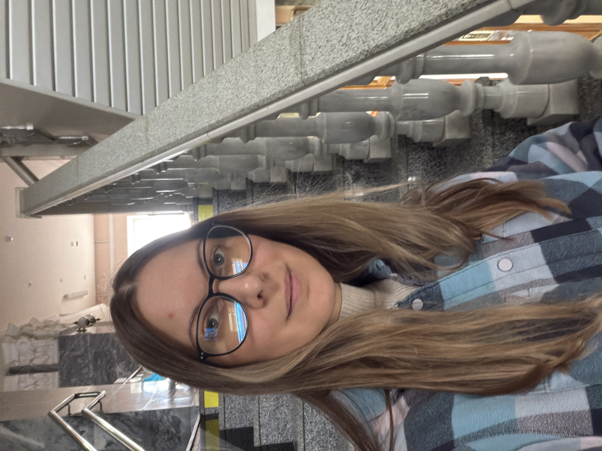 A young woman with long, straight light brown hair is captured in a close-up portrait, looking directly at the viewer with a calm, pleasant expression. She wears black-rimmed glasses, reflecting bright rectangular light sources, possibly windows or overhead fixtures. A small red blemish is visible on her forehead. Her attire consists of a light blue, white, and black plaid button-up shirt worn over a light cream or white turtleneck sweater. The shirt features white snap buttons.
The scene unfolds indoors, likely within a public or institutional building in Izhevsk, Russia, characterized by prominent architectural elements. The woman is positioned in front of a substantial staircase railing. This railing features a broad, textured light gray top rail, resembling granite or marble, supported by ornate, light gray balusters. Behind this, another, simpler metal railing with vertical slats is visible. The floor beneath appears to be composed of polished dark and light gray stone tiles. In the background, particularly to the upper left from the woman's perspective, a cream-colored wall and a brightly lit window are visible, suggesting daytime. Some areas behind her also show what might be construction materials or an unfinished section of the building. No visible text is present in the image.