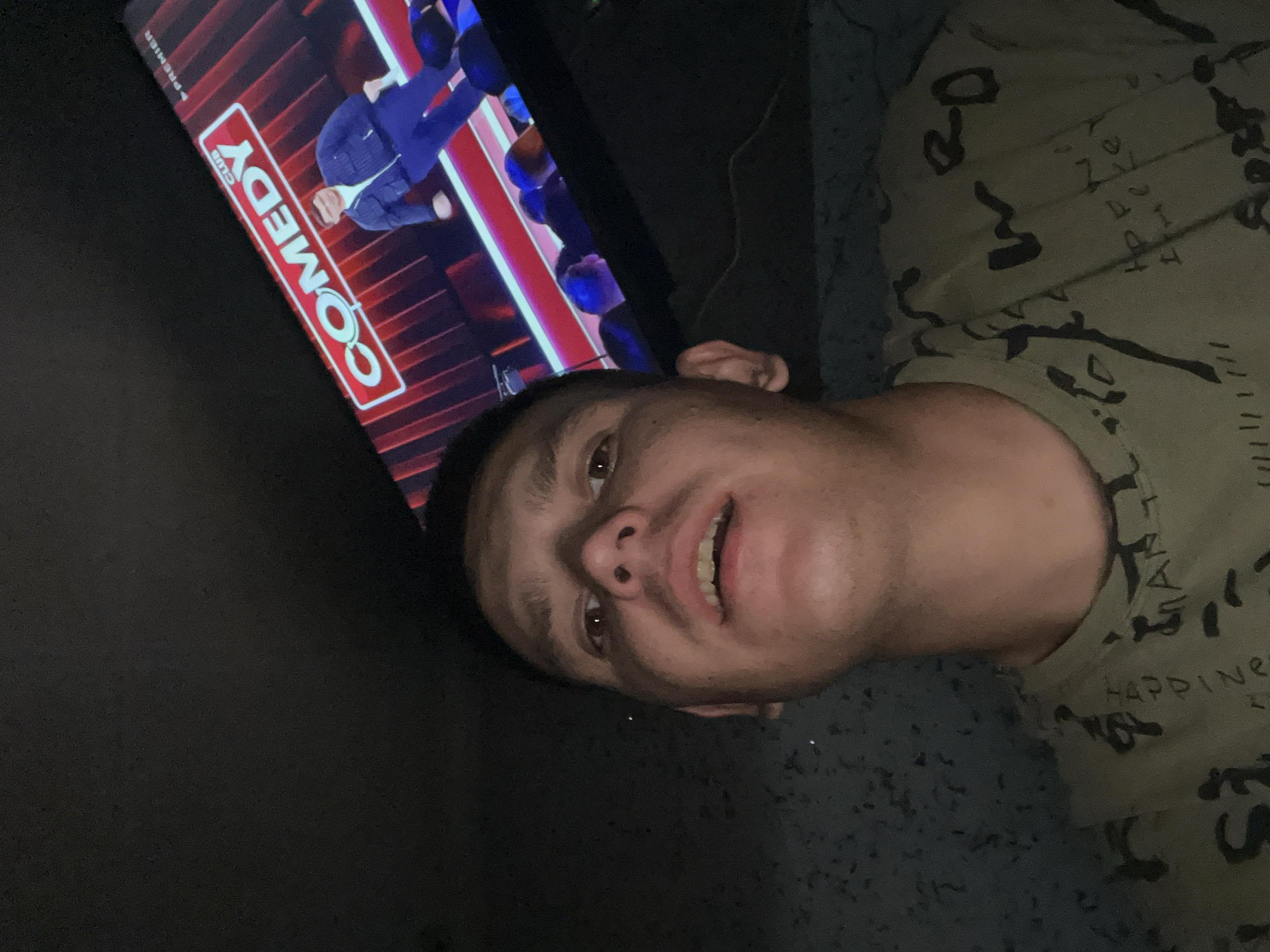 A low-angle, close-up selfie is taken by a man in what appears to be a dimly lit room. The man is looking directly at the camera with his mouth slightly open. He is wearing a light green t-shirt with a dark pattern of abstract shapes and what appears to be some text in a foreign language. In the background, a television screen is visible, displaying a comedy show with the word "COMEDY" prominently featured in large red and white lettering. A performer in a dark jacket and blue pants is standing on a stage in front of a red backdrop. The room is dark, with some speckled texture visible on the wall to the left of the man. The overall scene suggests a casual, indoor setting, possibly a living room, where the man is watching television. The specific location in the city of Дачное, Kyrgyzstan is not discernible from the visual information provided.