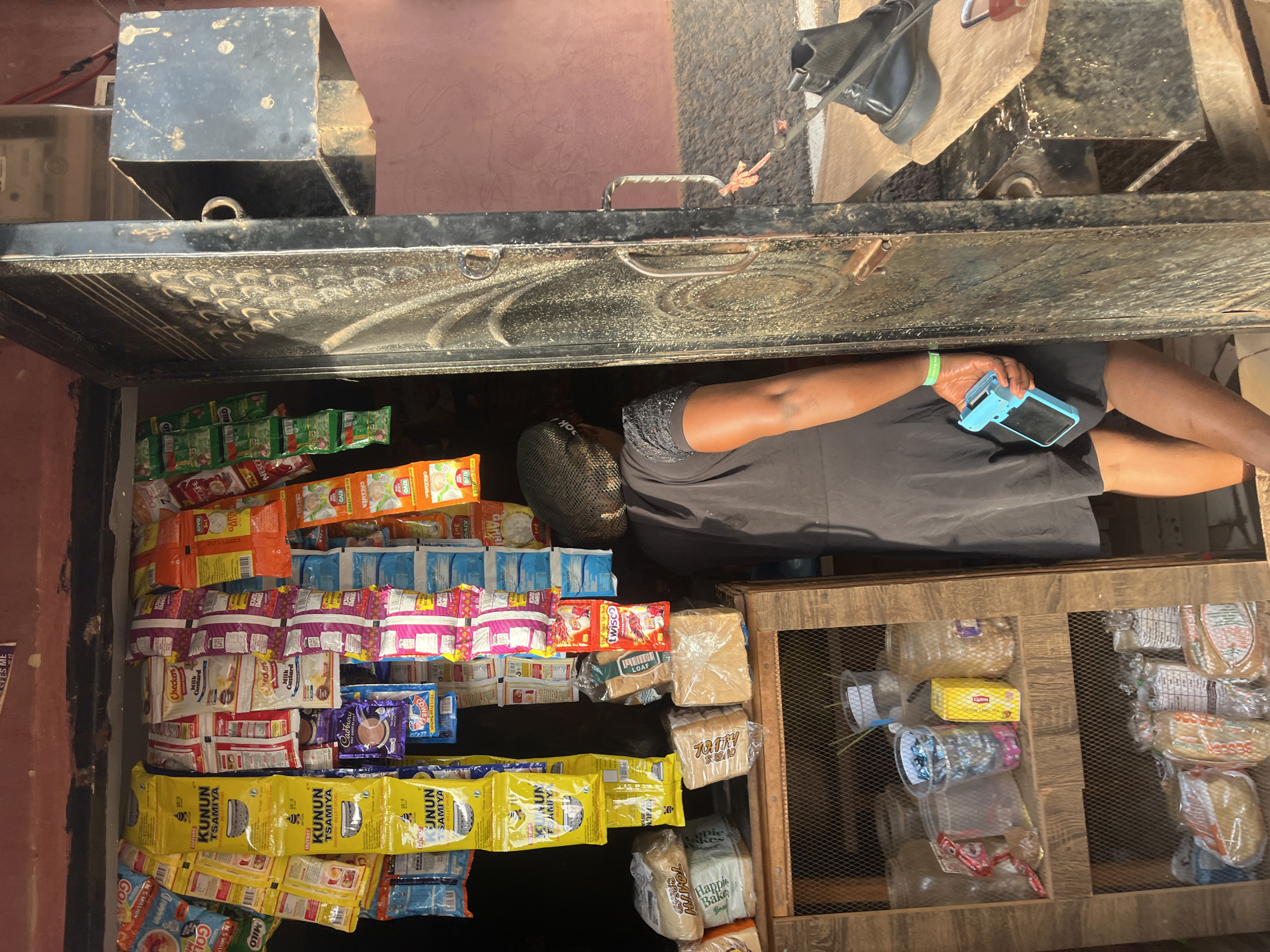 The image shows a woman looking at the goods displayed in a roadside shop in Abuja, Nigeria. The shop is a small, open-front structure with shelves stocked with various items, including packaged drinks, bread, and other food products. The shelves are densely packed with items such as Nestle products, Kunun Zaki, and different brands of bread. The woman is wearing a black shirt and holding a blue smartphone, likely browsing or taking a picture of the goods. The shopfront is metallic and hinged. Outside the shop, the ground is visible, and there are signs of urban activity.