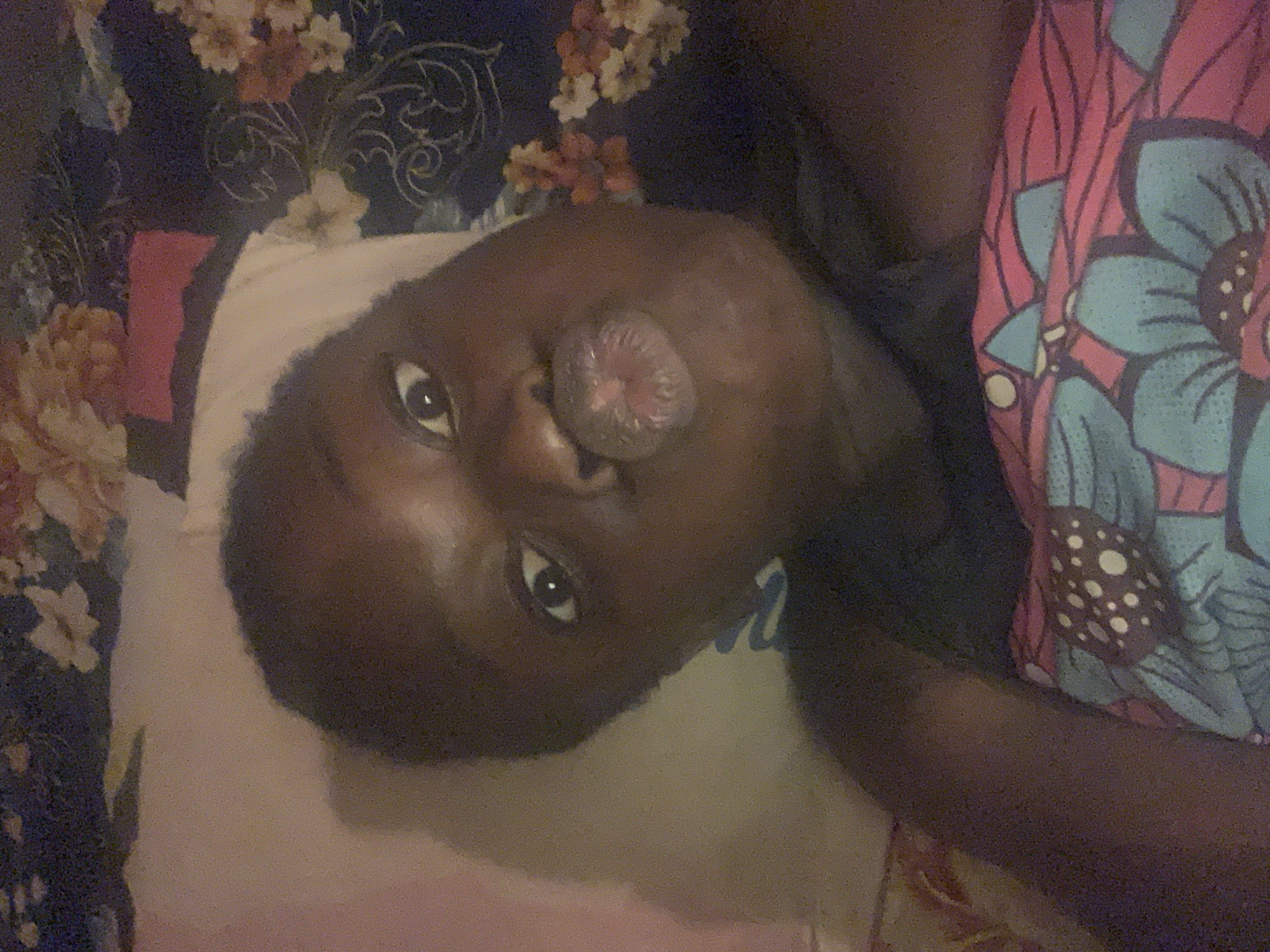 A dark-skinned person lies on a white pillow. The person's face is angled towards the camera. The lips are pursed in a kissing motion. The person wears a dark-colored shirt. A floral-patterned blanket is next to the person. A floral-patterned curtain is in the background. I am unable to determine the exact location of the scene in Agban, Nigeria, without further information.