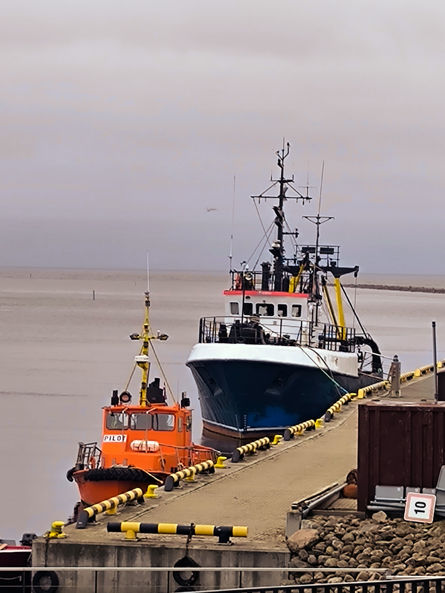 This is a medium shot of two boats docked at a pier in Salacgrīva, Latvia. The larger boat, a dark blue and white vessel, is positioned further back in the frame. In front of it, a smaller, bright orange boat with "PILOT" printed on its side is docked. The pier is made of concrete and lined with yellow and black bollards. There are rocks and a large brown container on the pier. The water is calm and the sky is overcast. In the distance, a small aircraft can be seen in the sky. There are no people clearly visible in the image.