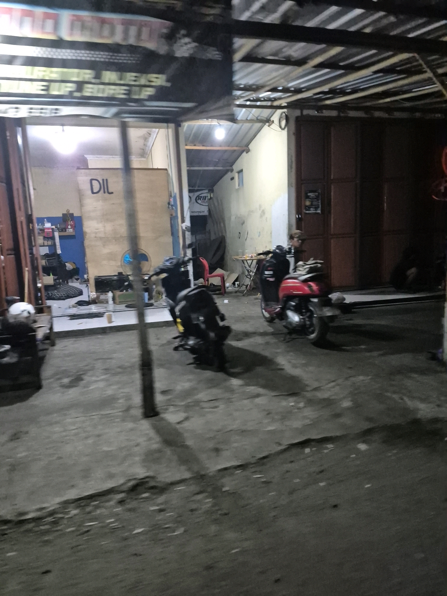In the city of Pabelan, Indonesia, an outdoor motorcycle repair shop is depicted at night. A sign above the entrance reads "MOTO". The shop appears to be cluttered with various items, including spare tires, tools, and a fan. Two motorcycles are parked outside the shop. One is a black motorcycle, and the other is a red scooter. The ground is paved with concrete and appears to be wet. The environment is dimly lit, suggesting it is late at night.