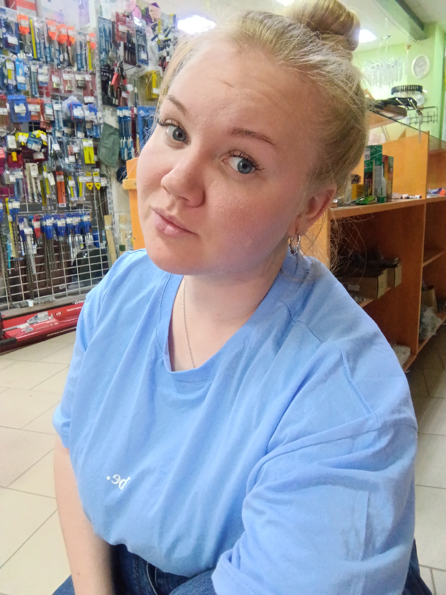 A blonde woman is posing for a photo in a hardware store. She has her hair in a bun and is wearing a blue t-shirt and silver earrings. She is leaning slightly to her left and giving the camera a slightly inquisitive look. In the background, there is a shelf stocked with hardware supplies and a wooden shelving unit. The store is well-lit, and the floor is tiled. It's mentioned that this picture was taken in the city of Фоки, Russia.
