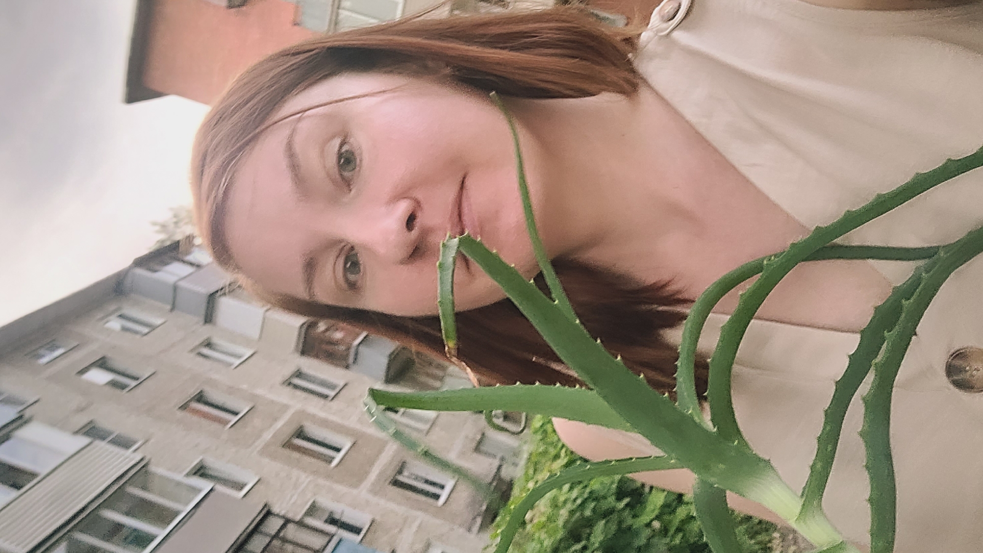 A woman with shoulder-length auburn hair holds a large aloe vera plant in front of her face, obscuring her mouth. She wears a light beige sleeveless top with two buttoned epaulets on her shoulders and a single button at the waist. The woman is positioned in the foreground, with an out-of-focus, multi-story residential building and some greenery in the background. The building appears to be made of concrete and has balconies and numerous windows. The sky above is overcast.