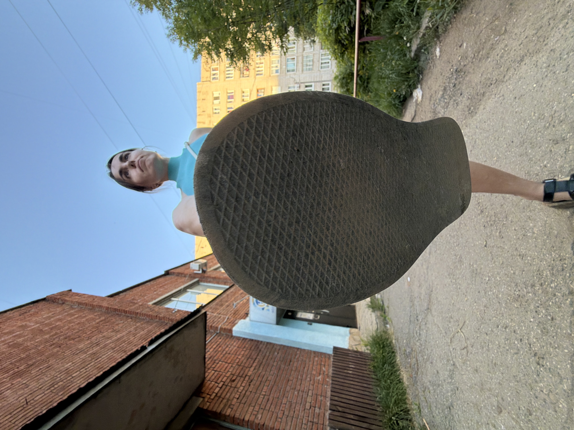 The photo depicts a view from a low angle looking upwards at a woman standing on a city street. The main focus is on the sole of her shoe, which is large and dark, dominating the foreground. Behind the sole, the woman is visible from the chest up, wearing a blue top. Her face is tilted slightly, and she looks down at the camera.
In the background, there are buildings on either side of the street. One is a red brick building, while another appears to be an apartment building with numerous windows. Vegetation is also visible along the roadside. The ground is paved with asphalt, and the sky is a clear blue.
Based on the location details provided, the photo was taken in the city of Izhevsk, Russia.