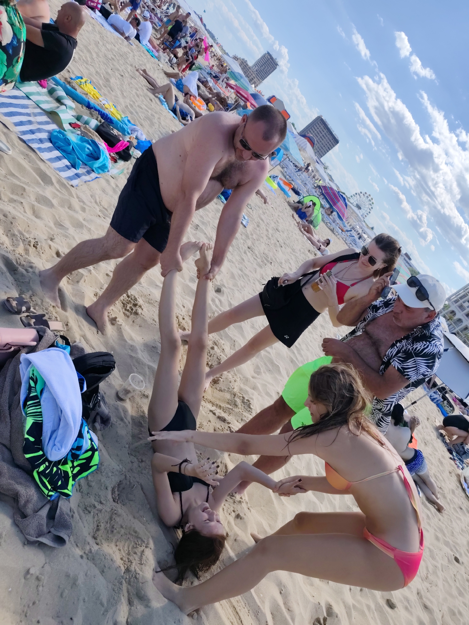 In Nesebar, Bulgaria, on a sandy beach, a group of people are engaged in various activities under a cloudy sky. In the foreground, a man in black shorts is assisting a young woman lying on her back, lifting her legs. She is wearing a black bikini. To her right, another young woman in a pink and orange bikini is sitting up, holding hands with the first woman. Further back, a man in a patterned shirt and white cap is standing and speaking with a woman in a red bikini and black skirt, who is holding a drink. In the background, the beach is crowded with people, beach chairs, umbrellas, and a Ferris wheel. The overall scene depicts a lively summer day at the beach.