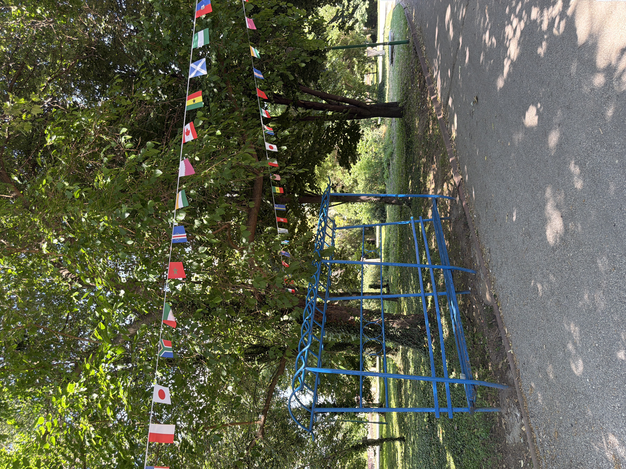 The image depicts an outdoor scene in a park-like setting. A string of international flags hangs across the frame, suspended from tree branches. Below the flags, a blue metal structure, possibly a shelter or seating area with benches, is visible. The ground is covered with gravel and foliage. Sunlight filters through the trees, casting dappled shadows on the path and ground. The scene appears to be set in a public area, likely a park or recreation ground. Specifically, this image was taken in the City of Pazardzhik, Bulgaria.