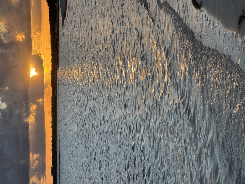 Golden sunset over water. The sun, partially obscured by clouds, casts a bright light with visible sunrays. The horizon line is defined by trees and utility poles silhouetted against the colorful sky. The water's surface reflects the sun's glow, creating streaks of golden light that contrast against the gray and blue water. The foreground shows the texture of the water, with small waves reflecting the evening light.