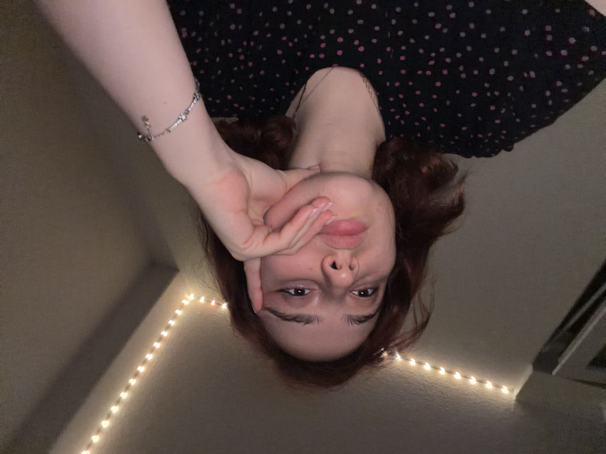 The image shows a young person with reddish-brown hair, resting their chin on their hand, looking towards the camera. They are wearing a black shirt with pink polka dots and a silver bracelet with charms. The background appears to be an indoor setting with a wall and a ceiling adorned with string lights. The lighting is dim, with the string lights creating a warm glow. The overall atmosphere is casual and personal. There is no information in the image that suggests a specific location in Strasbourg, France.