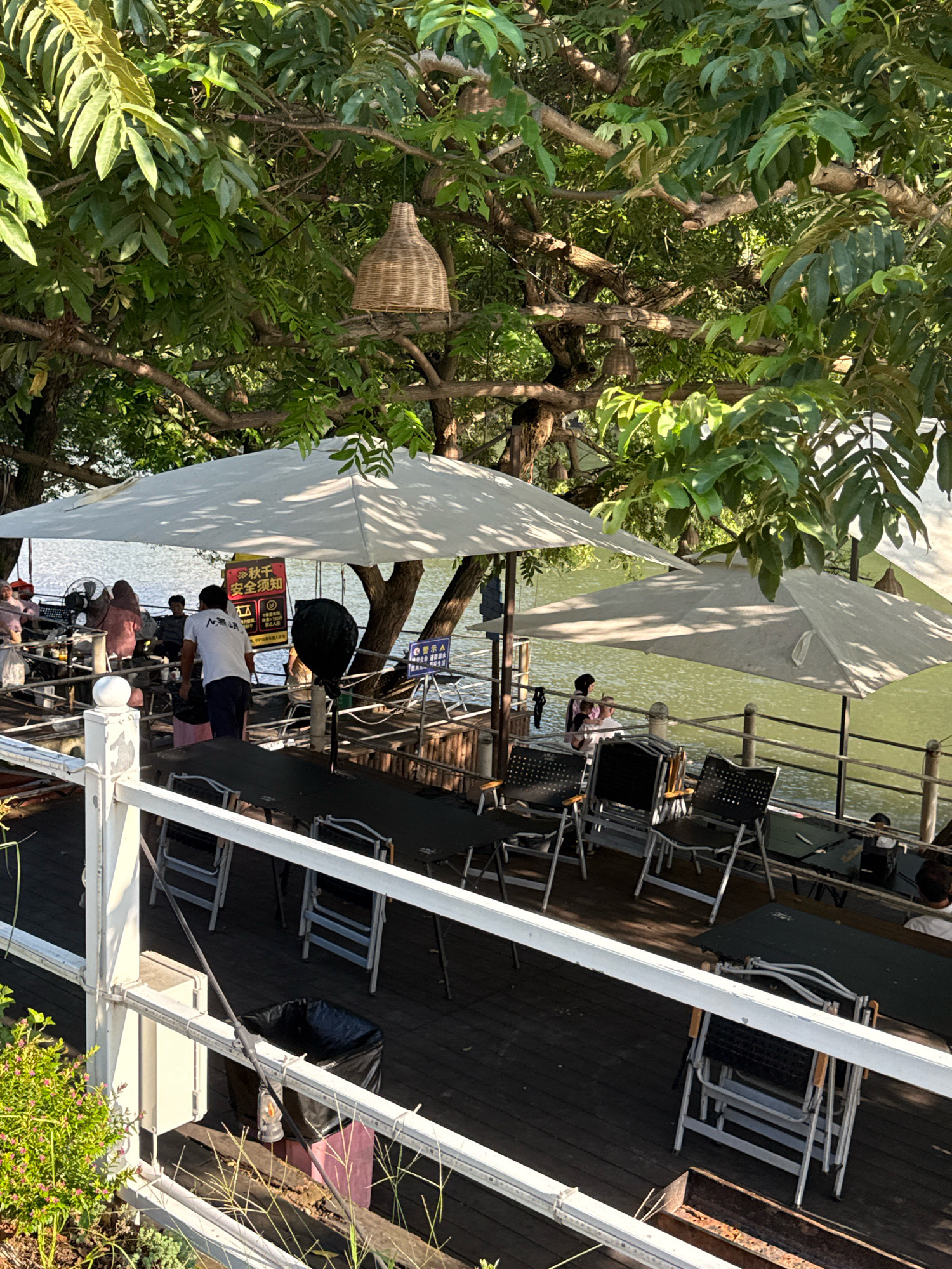 The image depicts an outdoor seating area adjacent to a body of water, likely a river or lake, in Yiwu, China. The scene is shaded by mature trees with lush green foliage, and a woven bamboo lamp hangs from a branch. The seating area features several tables and chairs, with a large white umbrella providing shade over a section of the tables. A few people are seated at tables, while others are standing or walking. One man in a white shirt is visible from behind, facing away from the camera. Another person, partially obscured, is walking towards the water. There's a prominent black trash can on the wooden deck. In the background, the water is calm and reflects the surrounding greenery. A red and yellow sign with Chinese characters is posted on a pole near the tables. The overall impression is a relaxed, outdoor dining or gathering space.