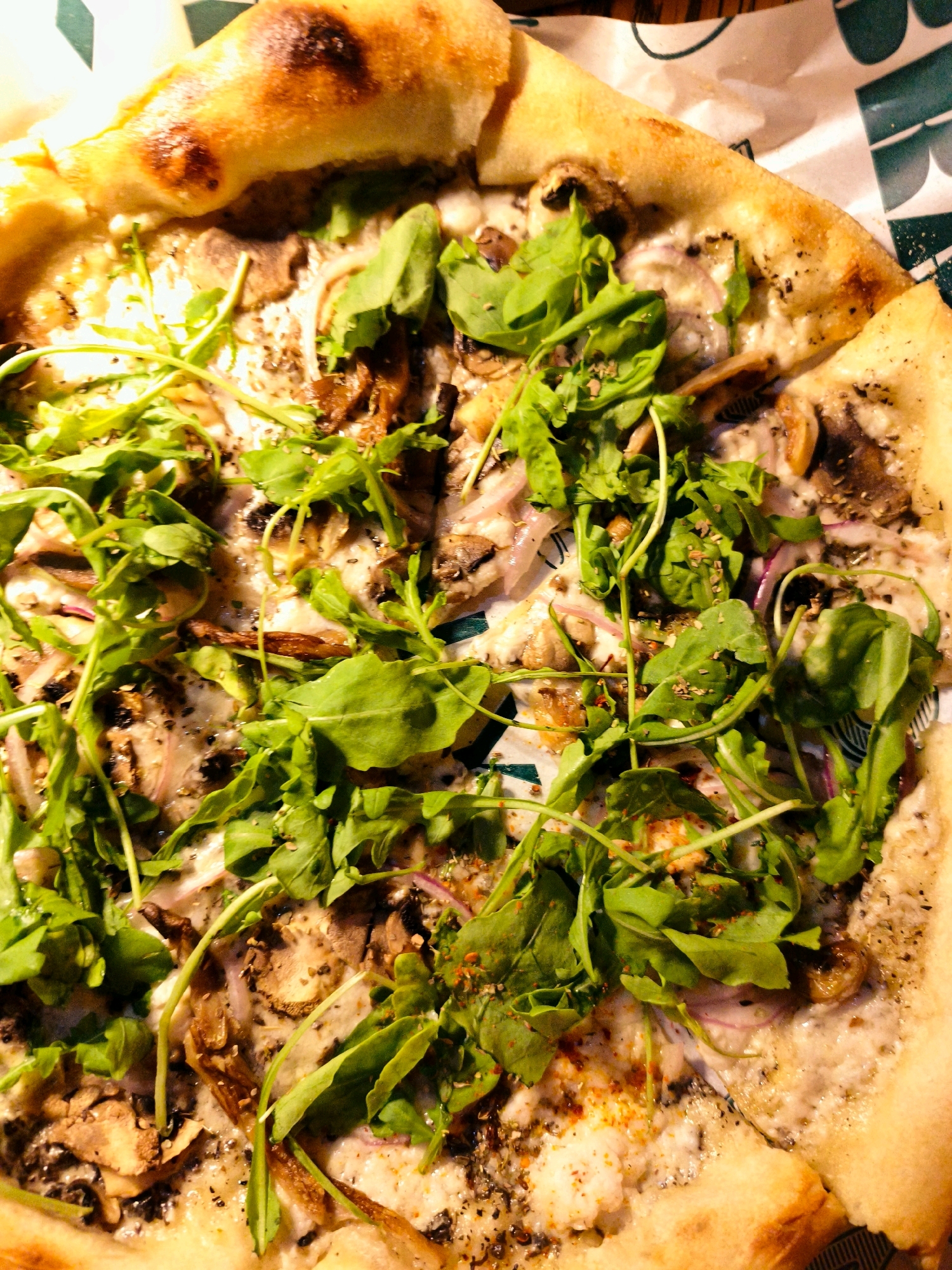 A close-up, overhead shot of a cooked pizza is presented, with the crust prominently featured on the left and top edges. The pizza is topped with melted cheese, sliced mushrooms, thin slivers of red onion, and a generous scattering of fresh green arugula leaves. Dried herbs and spices, including what appears to be red pepper flakes and black pepper, are visible on the cheese and crust. The pizza is resting on a piece of patterned paper, which appears to be part of its packaging. The background shows a glimpse of dark green and white patterned paper, possibly from the same packaging. The lighting is warm and artificial, suggesting an indoor setting. There are no people or animals present in the image. Based on visual cues alone, it is not possible to determine the specific location in Be'er Sheva, Israel.