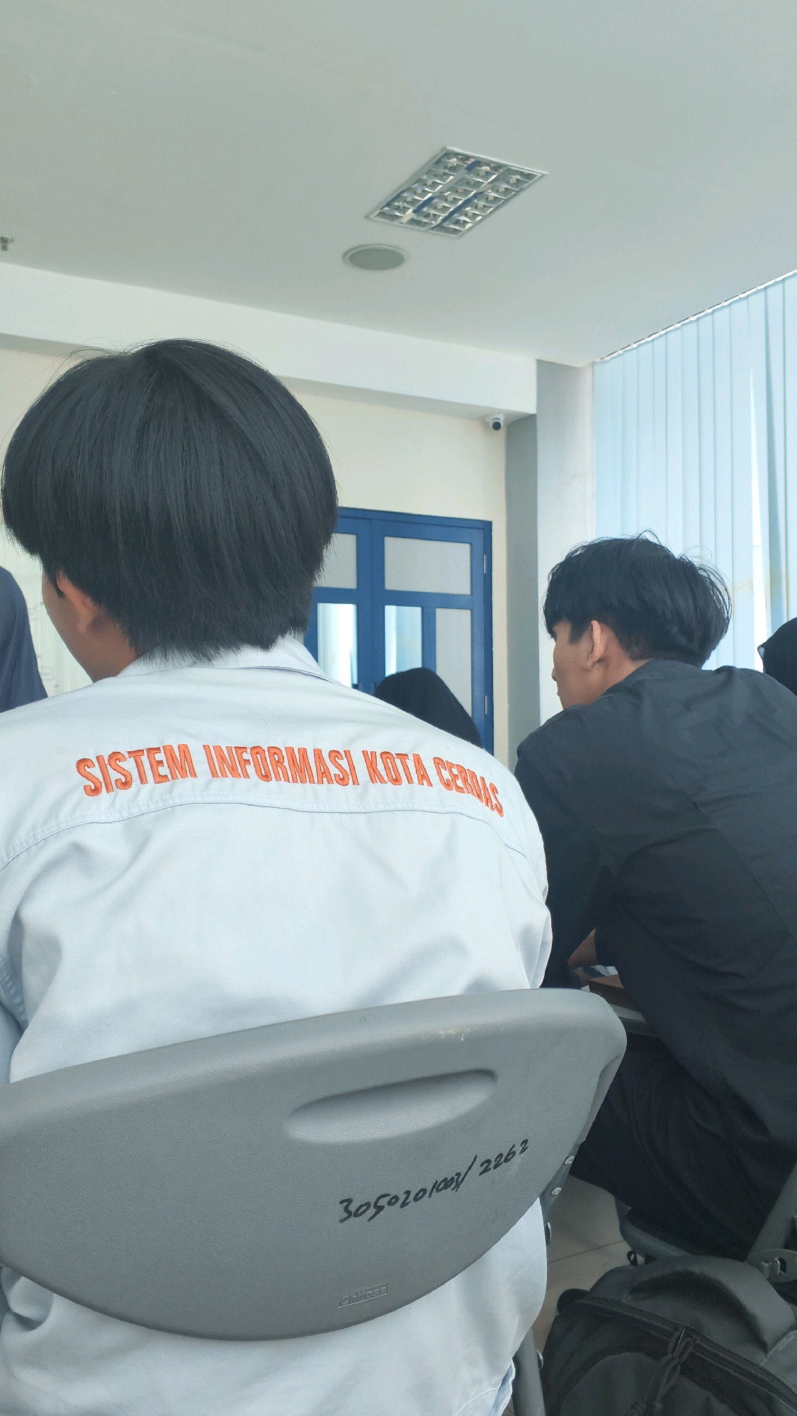 The image shows the back of two young men sitting in a classroom or meeting room. The man in the foreground is wearing a light-colored shirt with orange text that reads "SISTEM INFORMASI KOTA CERDAS". The man behind him is wearing a black shirt. A grey chair with writing on it is in the foreground. In the background, there is a blue door and a window with vertical blinds. The ceiling has a fluorescent light fixture and a circular speaker. There is no information in the image that indicates the location is Lohbener, Indonesia.