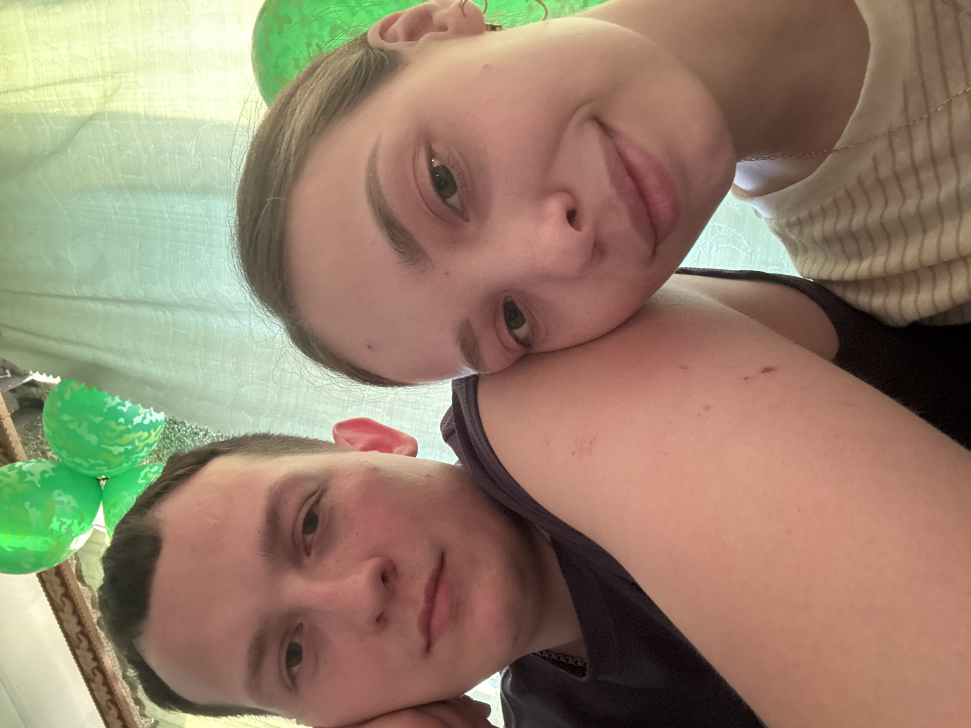A selfie photograph shows a young adult female with fair skin resting her head on the arm of a young adult male with fair skin. The female has light brown hair pulled back, visible earrings, and a striped shirt. The male has dark hair and is wearing a dark-colored shirt and a chain necklace. Green balloons are visible in the background. The location is inferred from the prompt to be in Кемуль, Russia, though no evidence from the image itself supports this.