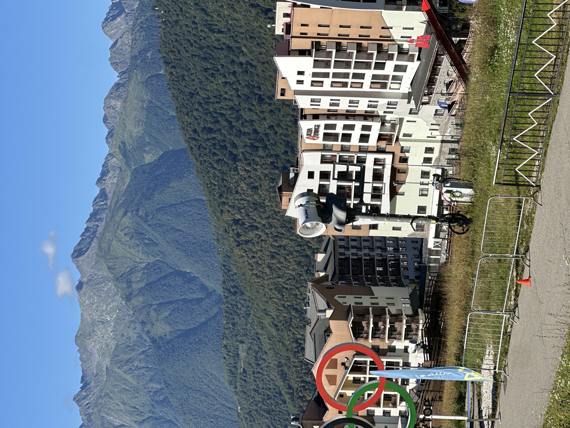 The image depicts a mountainous landscape with buildings in the foreground, situated in Estosadok, Russia. The Olympic rings are prominently displayed on the left side of the frame. Several multi-story buildings with modern architecture, featuring balconies and large windows, are clustered together. In the center, a tall pole with a large fan-like attachment is visible, possibly a snow cannon or fan. A blue flag is planted near the Olympic rings. The area is surrounded by lush green mountains, with a clear blue sky overhead. A paved path and a metal fence are in the foreground, with some grass and wildflowers. A bicycle is parked next to the pole with the fan attachment. The scene suggests a resort or sporting venue in a mountainous region.