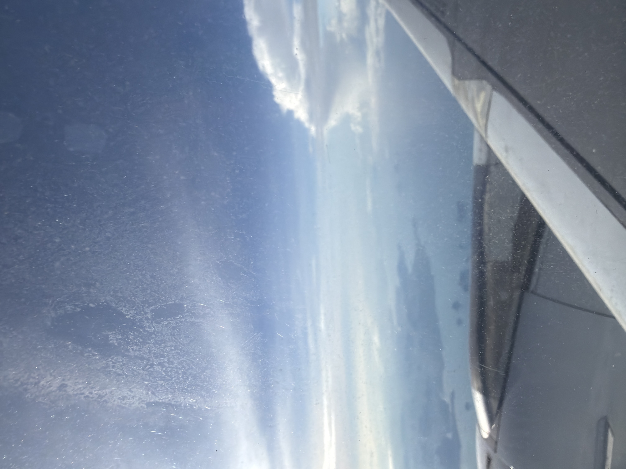 The image is a photograph taken from an airplane window, looking out at the sky and the ground below. The sky is a bright blue with scattered clouds. The ground appears to be a landscape of islands and water, possibly the coast of Thailand. The airplane wing is visible in the foreground, and the window has some smudges and streaks on it. The image is taken from a high altitude, and the light is bright, suggesting it is daytime. The location is Sisa Chorakhe Noi Subdistrict, Thailand.