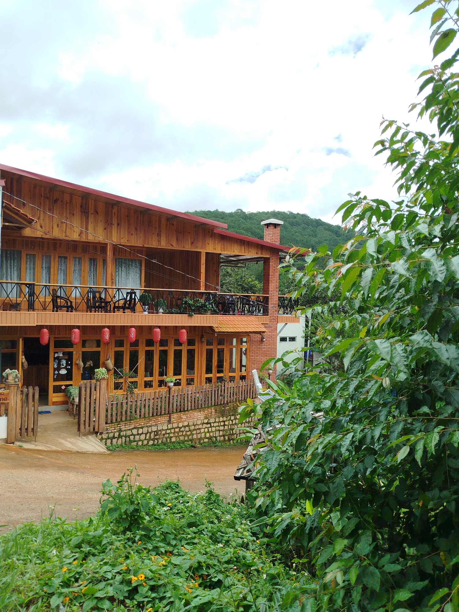 The image shows a two-story wooden building with a red tiled roof and a brick chimney, situated in a rural or mountainous area of Da Lat, Vietnam. The building has large windows and balconies, with red lanterns hanging from the lower balcony. In the foreground, there are lush green plants and small yellow flowers. The background features a forested hillside under a cloudy sky. The scene appears to be a cafe, guesthouse, or restaurant, possibly located on a scenic route outside the city center of Da Lat, known for its cooler climate, pine forests, and French colonial architecture.