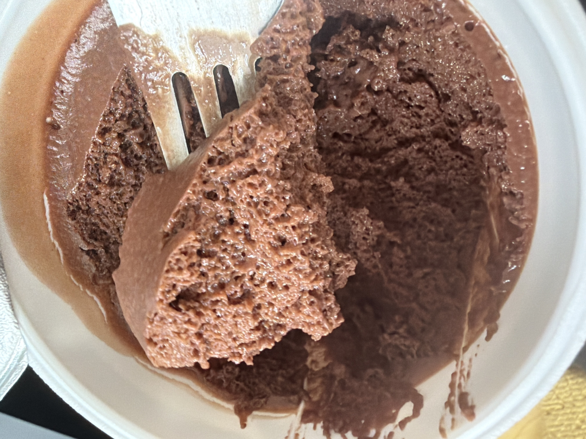 A close-up, slightly angled shot shows a white container filled with chocolate mousse, partially consumed. A silver fork is embedded in the mousse, holding a large piece with a porous texture. The remaining mousse appears smooth. The container shows signs of melting, with a thin layer of liquid surrounding the mousse. No people are visible. The location is not discernible from the image.