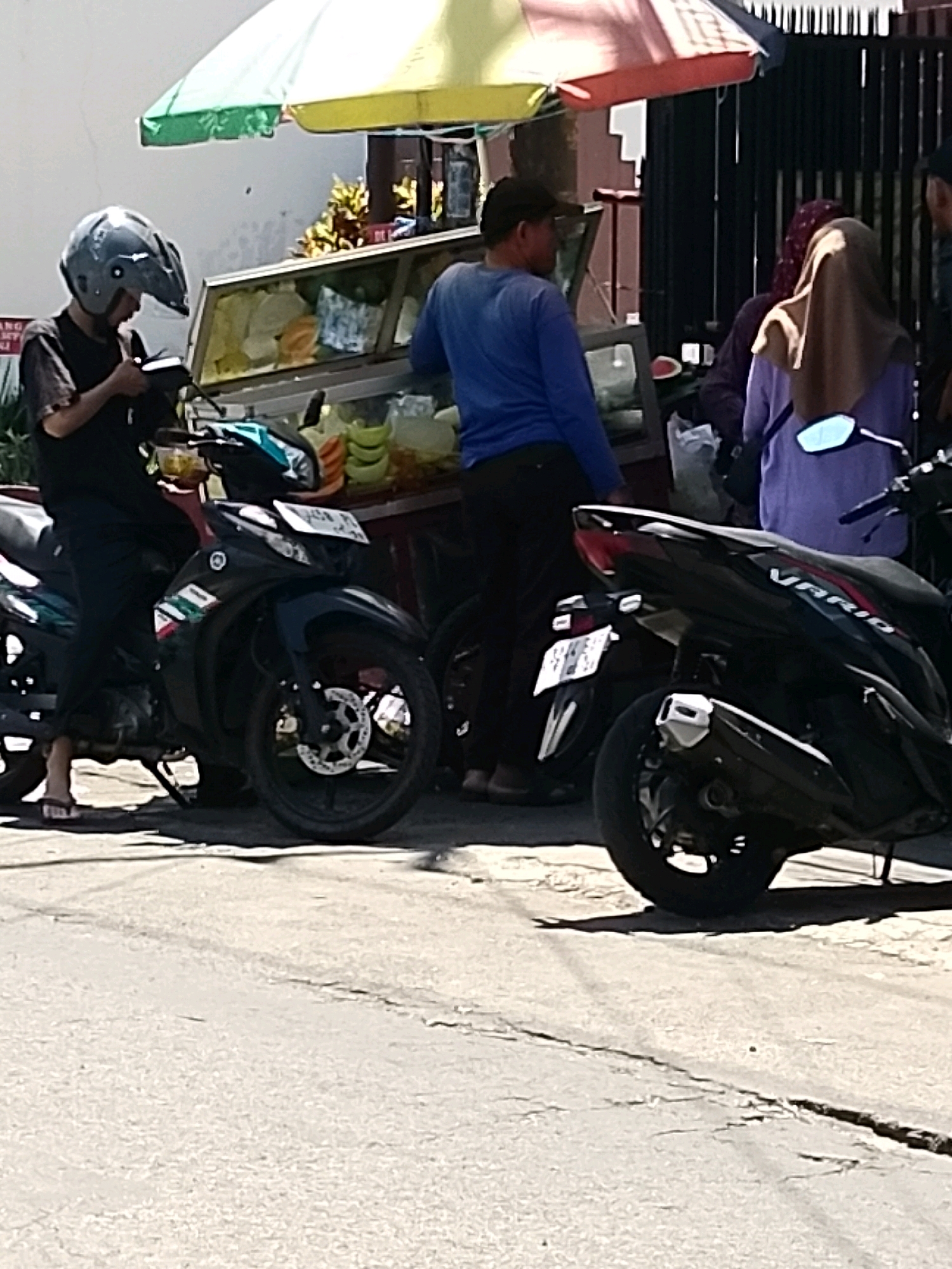 The image depicts a street scene in Surabaya, Indonesia, with a focus on several individuals and motorcycles. In the foreground, a person wearing a black t-shirt and dark pants is seated on a black motorcycle. They are wearing a grey helmet and appear to be looking at their phone. The motorcycle has a license plate that is partially visible. To the right of this motorcycle, another black motorcycle is parked, partially obscuring the view of the street.
Behind the motorcycles, a food stall is set up under a colorful awning. The stall contains a display of food items, including what appear to be fruits and vegetables in clear containers. Two individuals are visible near the food stall. One person is wearing a blue shirt and dark pants, standing and looking towards the stall. Another person, wearing a hijab and a light purple top, is also near the stall, with their back to the camera.
The environment suggests an outdoor commercial setting, likely a street market or roadside food vendor area. The pavement appears to be asphalt, with some cracks visible. The lighting indicates it is daytime. The overall impression is a snapshot of everyday life in a busy urban environment.