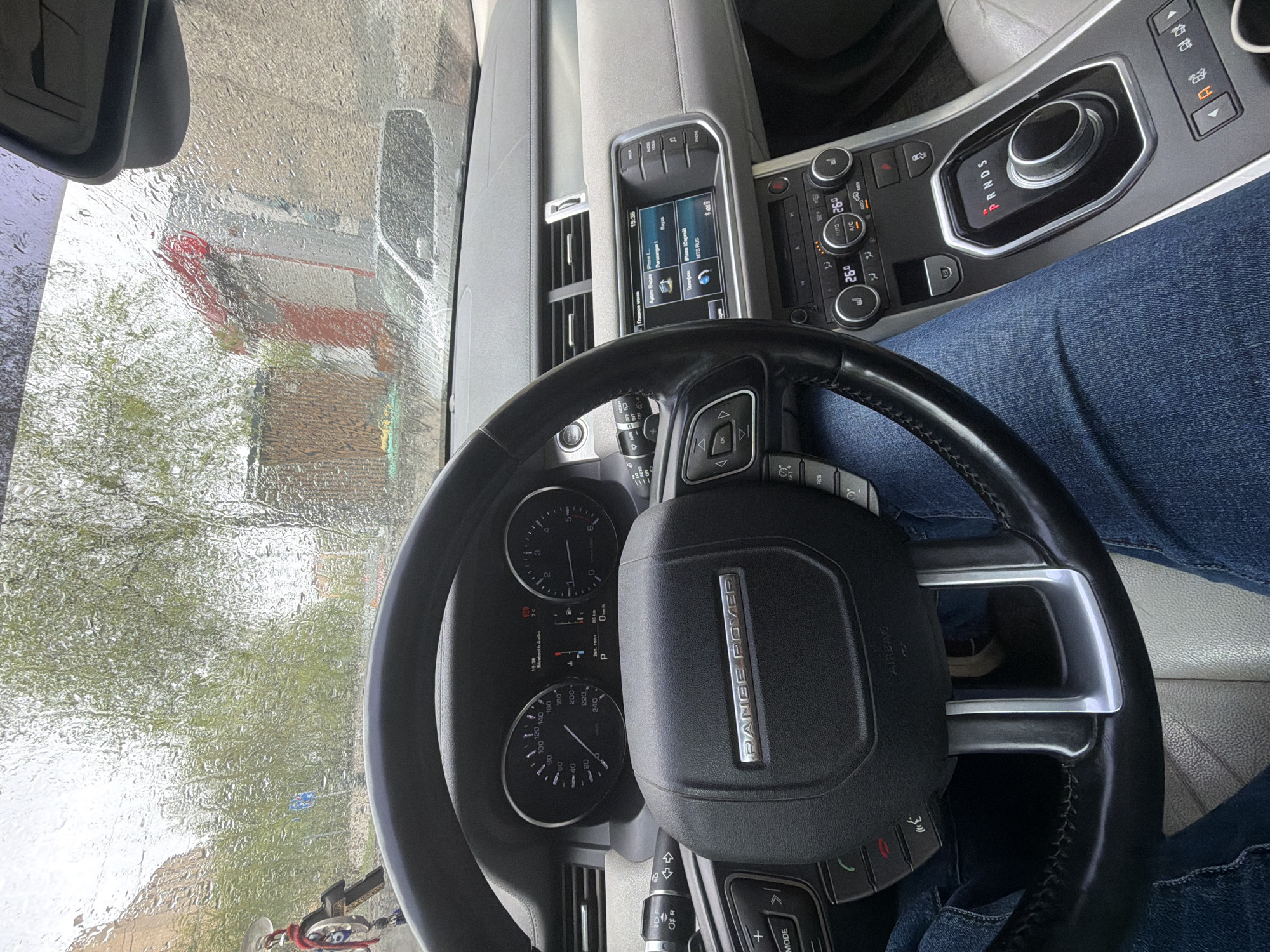 The image shows the interior of a Range Rover vehicle, including the dashboard, steering wheel, and a glimpse of the driver's denim-clad legs. The car's console displays the shift lever in Drive (D). The car is stationary, as indicated by the speedometer at 0 km/h. The infotainment screen is active, showing what appears to be a phone interface. The view through the windshield shows a rainy day with blurred visibility of the exterior surroundings. Based on the presence of Russian language on the infotainment screen (e.g., "Главное," "Телефон," "МТС RUS") and the OCR detected text, the car's location is in Russia. Given the context of the document, it is likely that the car is in the city of Krasnoyarsk.