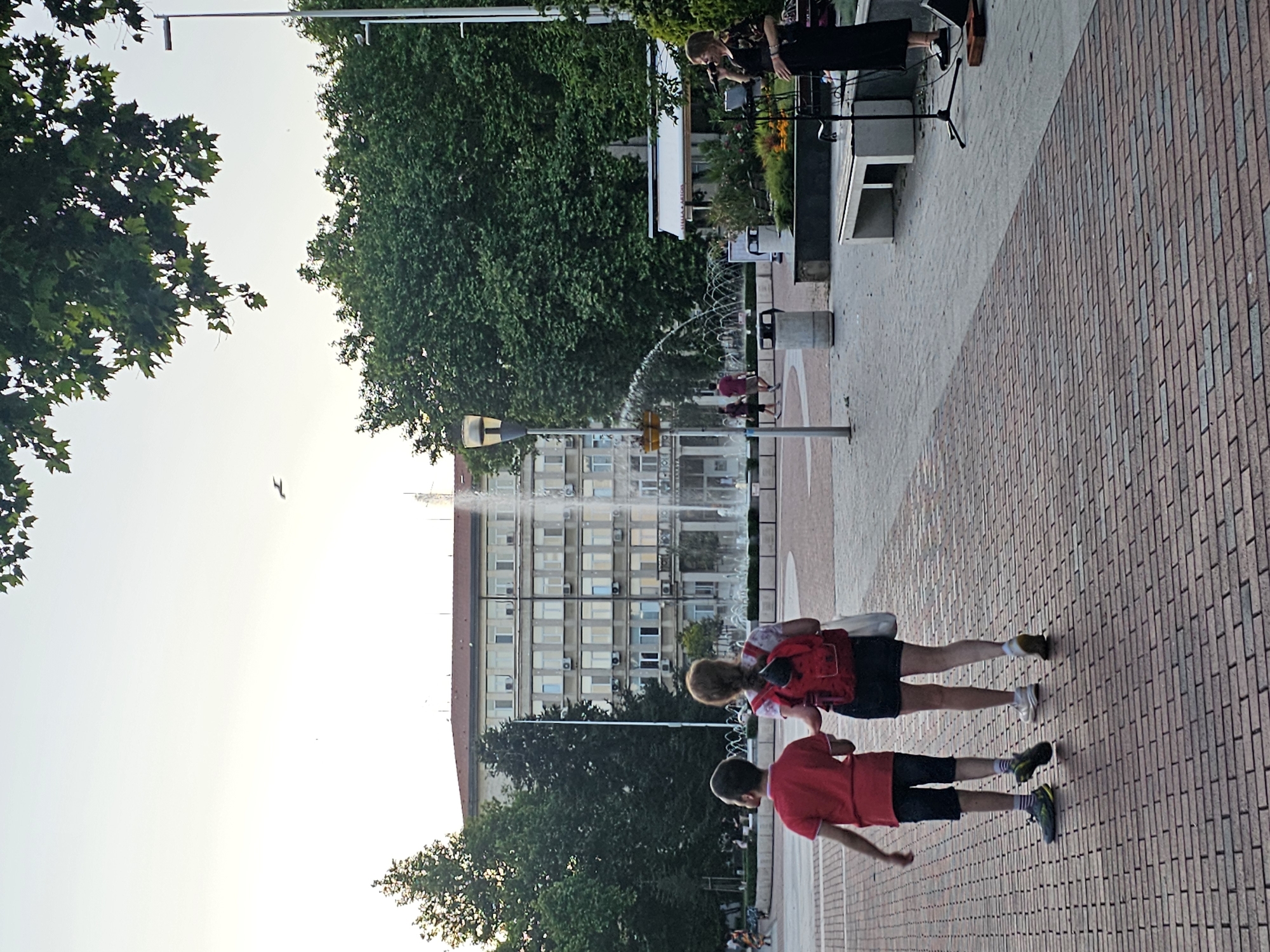 In a city square, two children, a boy in a red shirt and shorts and a girl with a red backpack and shorts, walk away from the camera on a brick-patterned walkway. In the background, a fountain sprays water in front of a large, multi-story building with many windows. To the right, a woman in a black dress sings into a microphone on a small stage. Trees and streetlights are visible throughout the scene.