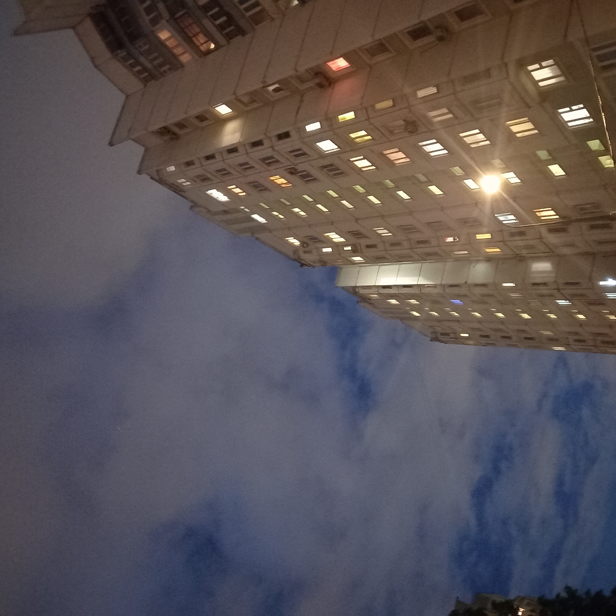 Here is a detailed description of the image:
The photograph presents a low-angle shot of two large apartment buildings at night. The buildings are illuminated with light emanating from numerous windows. The architectural style is typical of Soviet-era residential construction, with repetitive patterns of rectangular windows and a generally boxy structure.
The sky is a prominent feature, occupying a significant portion of the frame. It is filled with dark, swirling clouds, with patches of blue visible in between. The overall ambiance is slightly ominous and atmospheric.
A street lamp is positioned near the center of the buildings, casting a bright glow that illuminates the surrounding windows. The angle and perspective suggest that the photo was taken from street level, looking upwards towards the buildings.
While the image does not contain people, the presence of lights in the windows indicates occupancy within the buildings. The scene is set in an urban environment, likely a residential area in a city. Based on architectural style, this image is most likely in Moscow, Russia.