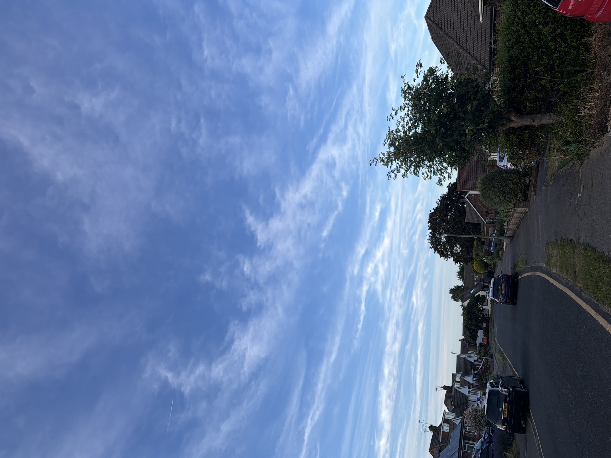 The image depicts a residential street scene in Maidstone, United Kingdom, under a bright blue sky with scattered white cirrus and altocumulus clouds. A faint aircraft contrail is visible in the upper-left portion of the sky. The foreground on the right features a paved sidewalk with overgrown grass and weeds, leading to a small, leafy green tree partially obscuring a red vehicle. A trimmed green hedge runs along the property line behind the tree. The asphalt road in the mid-ground curves gently to the right and features a faded yellow line along its right edge. Two vehicles are prominent on the road: a dark-colored Toyota Verso (registration DG59 AOZ) parked on the left side facing away from the viewer, and a dark-colored SUV (registration DG69 RCZ) parked on the right side, also facing away. Along both sides of the street, residential houses, primarily detached or semi-detached, are visible with pitched, tiled roofs; some exhibit solar panel installations. Gardens, hedges, and various trees, including a large, dark-foliaged tree on the right, line the properties. A street lamp post is visible on the right side of the road in the mid-ground. No individuals are present in the scene. The overall lighting suggests late afternoon or early evening conditions.