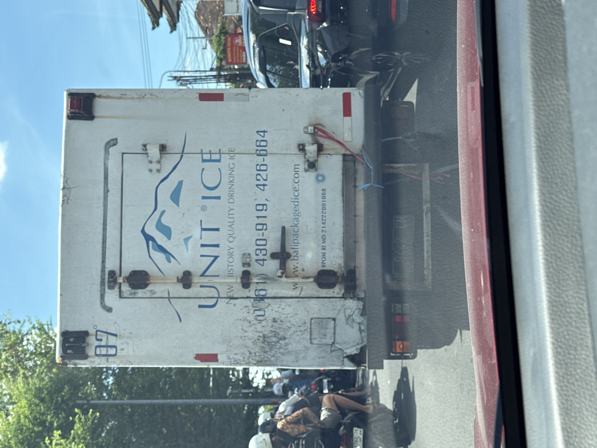 The image depicts the back of a white delivery truck for "Unit Ice," a company specializing in drinking ice, captured in what appears to be Kuta, Indonesia. The truck's signage includes phone numbers and a website address. The truck is situated in traffic alongside a black car. A red vehicle frames the right side of the shot, suggesting the photo was taken from inside another car. On the left side, a portion of a tree and a streetlight are visible, along with two people on a motorbike. The license plate number 8442 is visible on the truck.