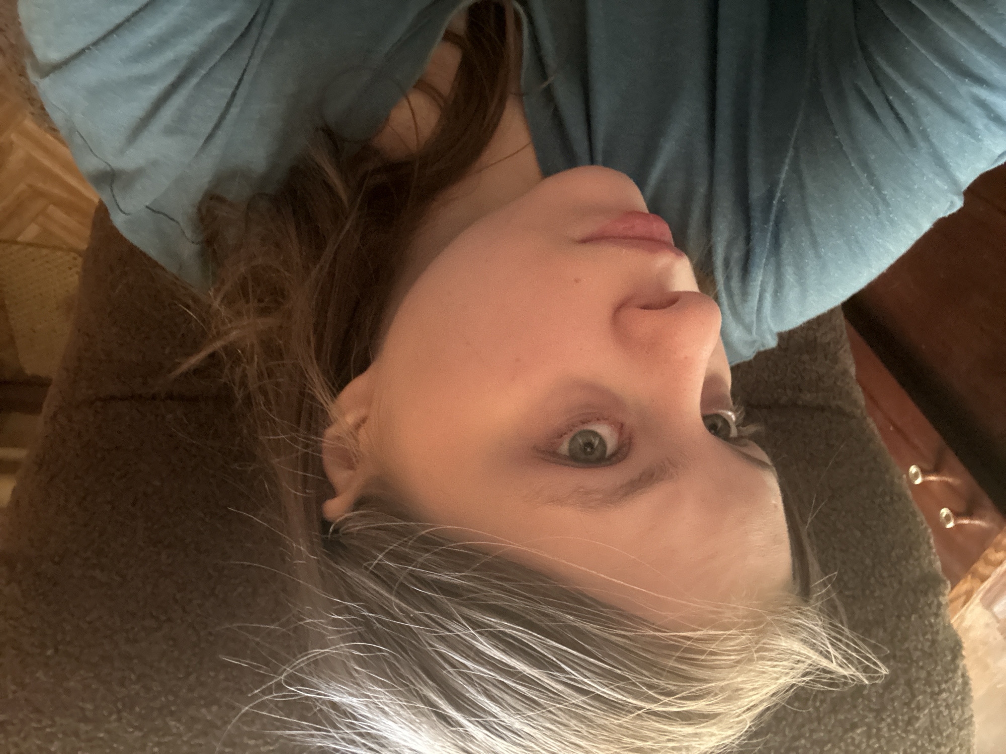 A Caucasian woman with green eyes and graying blonde hair is positioned in the center of the image, wearing a blue shirt. She appears to be lying on a brown textured surface, possibly a couch or a rug. The background includes a wooden floor and a wooden dresser with metal drawer pulls. The location is not Moscow, Russia, and cannot be determined from the image.