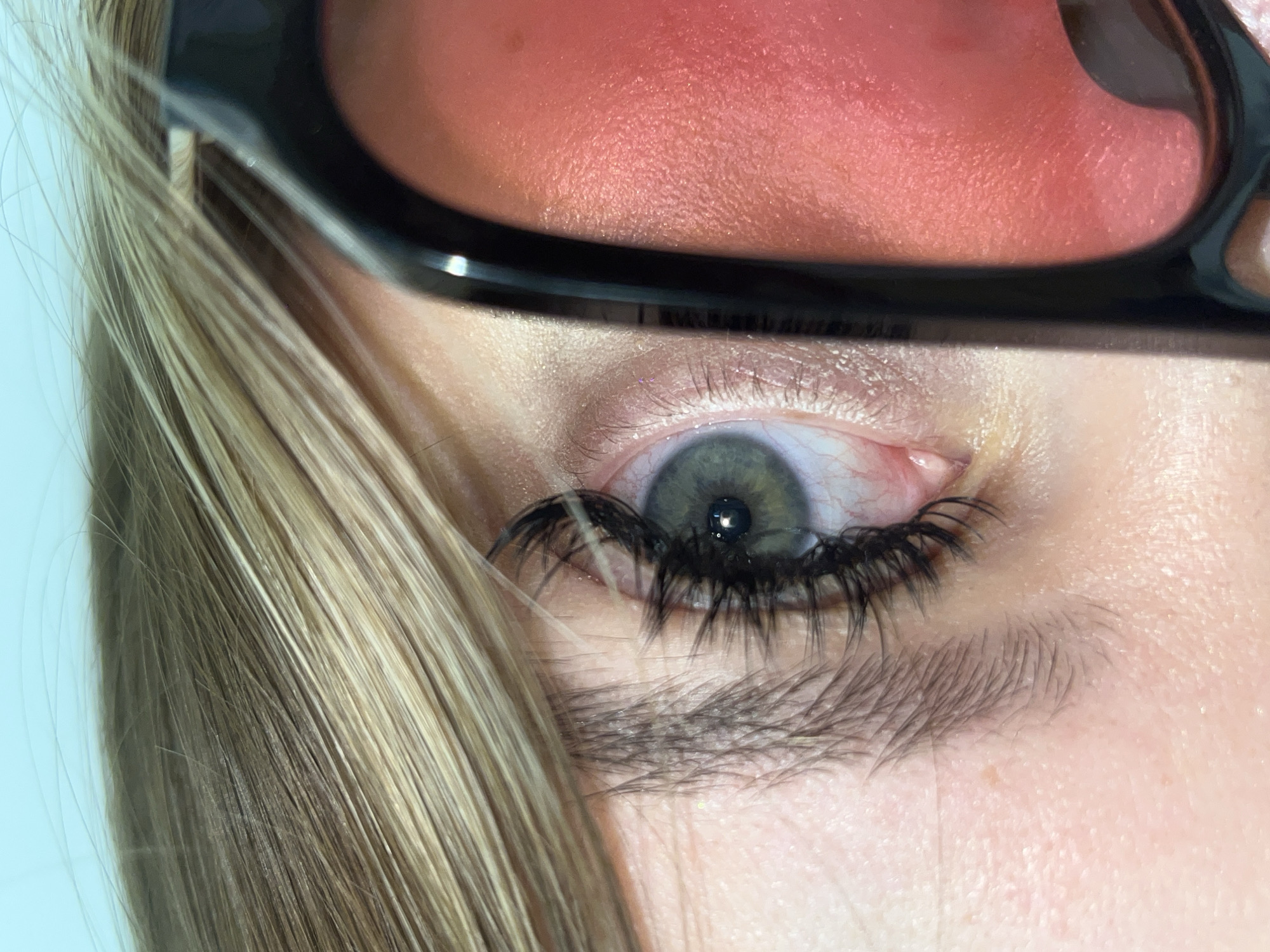 This close-up, high-angle shot centers on the left eye and surrounding facial features of a person, reportedly located in Krasnodar, Russia. The composition is tightly framed, focusing on the eye, artificial eyelashes, and a portion of sunglasses and hair.
The subject's eye features a distinct grey-green iris with a clearly visible black pupil, reflecting a small light source. Long, voluminous, and dark artificial eyelashes dramatically frame the upper eyelid, extending outward. Below the eye, the natural eyebrow is dark and appears relatively thick, though partially obscured.
A prominent pair of sunglasses, with thick black frames and red-tinted lenses, covers the upper part of the face, casting a reddish glow over the skin of the brow area. The sunglass frame is visible across the top of the eye. Light blonde or light brown hair, appearing straight and fine, flows from the left side of the frame, partially covering the temple and obscuring the sunglass arm. The skin around the eye shows texture with visible pores and fine facial hairs.
No specific activity is explicitly depicted, and the person's expression cannot be discerned from this cropped view. The lighting is bright, creating clear highlights on the skin and the sunglasses. There is no visible text in the image. The background is a plain, light color, suggesting an indoor setting.
