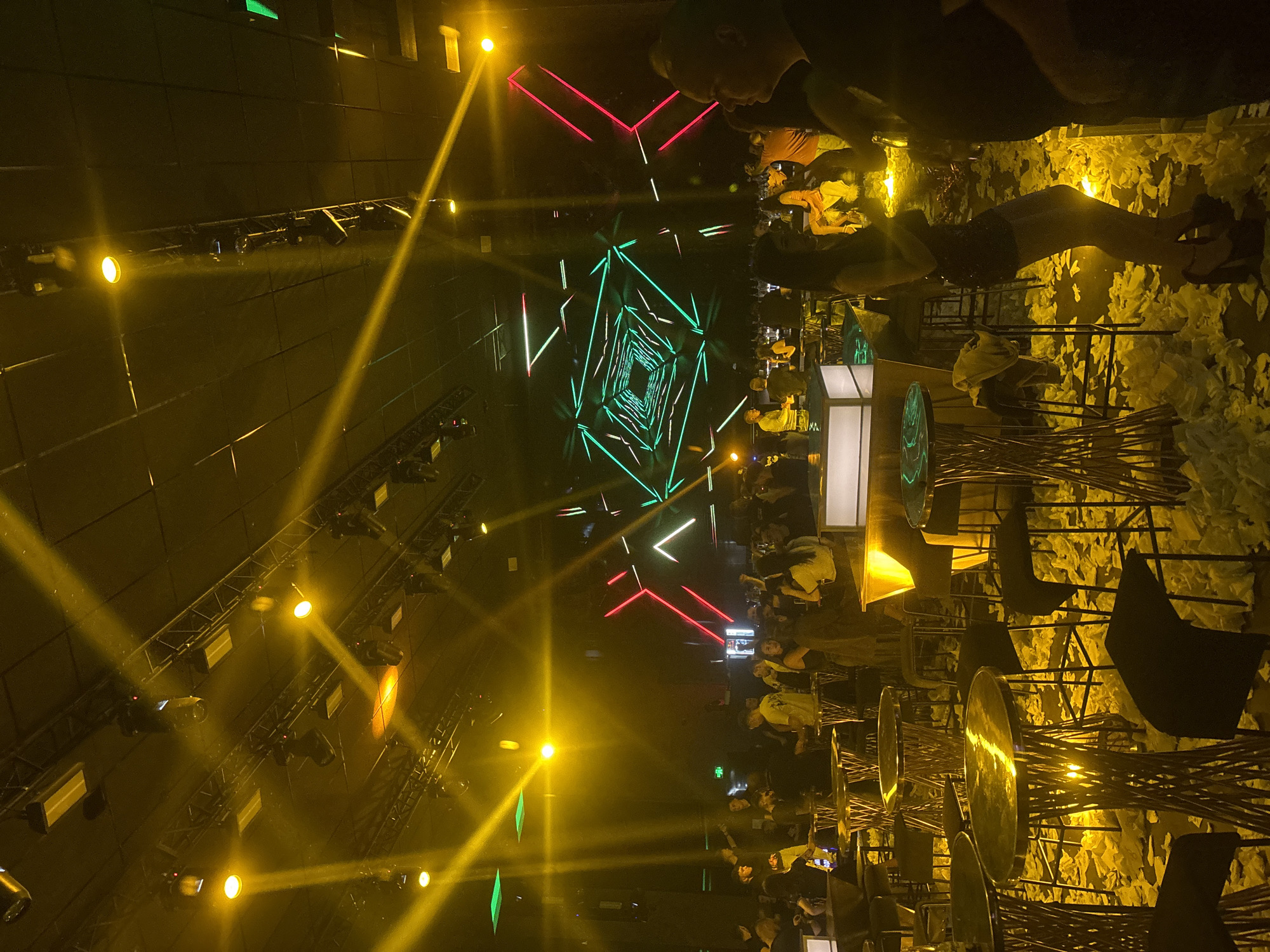 The image depicts an indoor event space, possibly a nightclub or concert venue, in Stara Zagora, Bulgaria. The scene is dominated by a complex lighting system overhead, featuring numerous yellow spotlights, and green and red neon geometric shapes. A crowd of people is present, some seated at high-top tables with metal chairs. The tables appear to have reflective surfaces. The background includes wall decorations possibly resembling foliage.